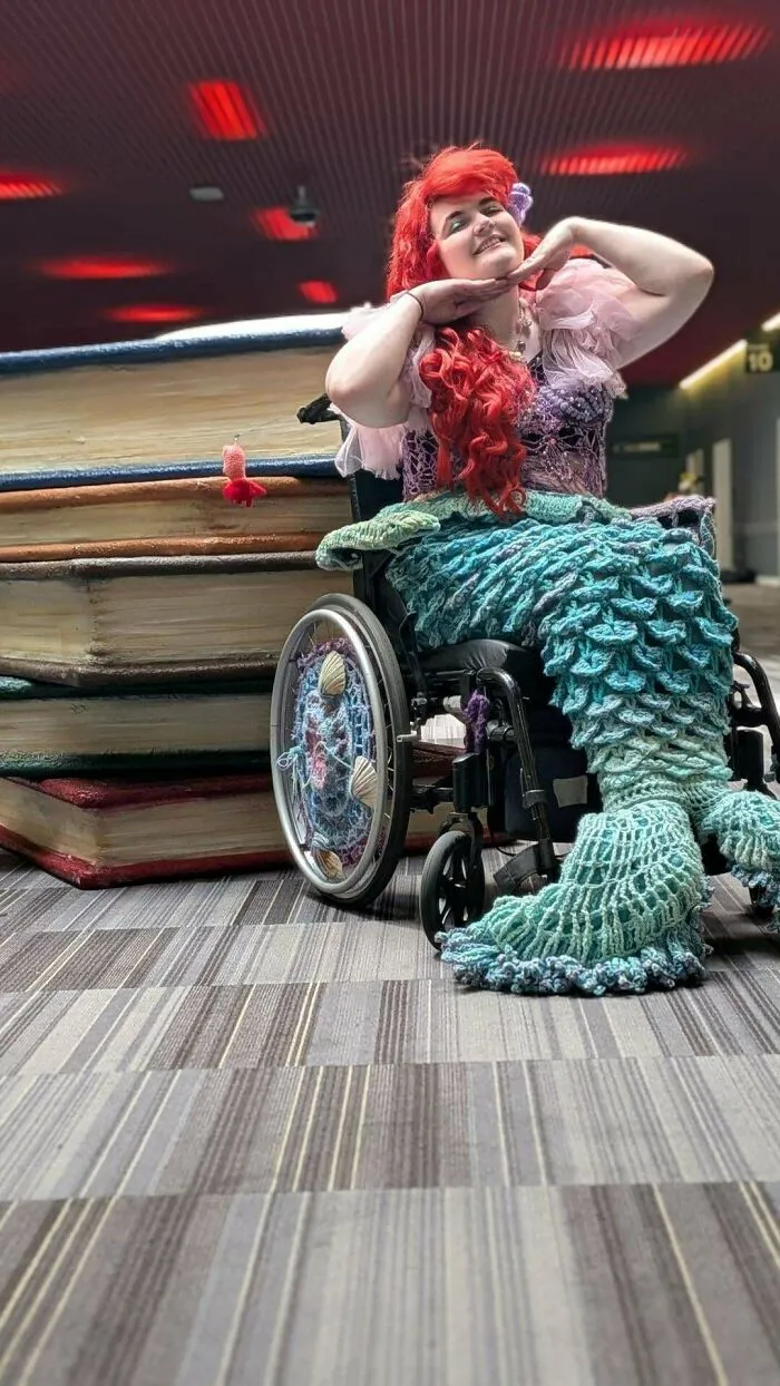 Person dressed as a crocheted mermaid in wheelchair next to giant stacked books