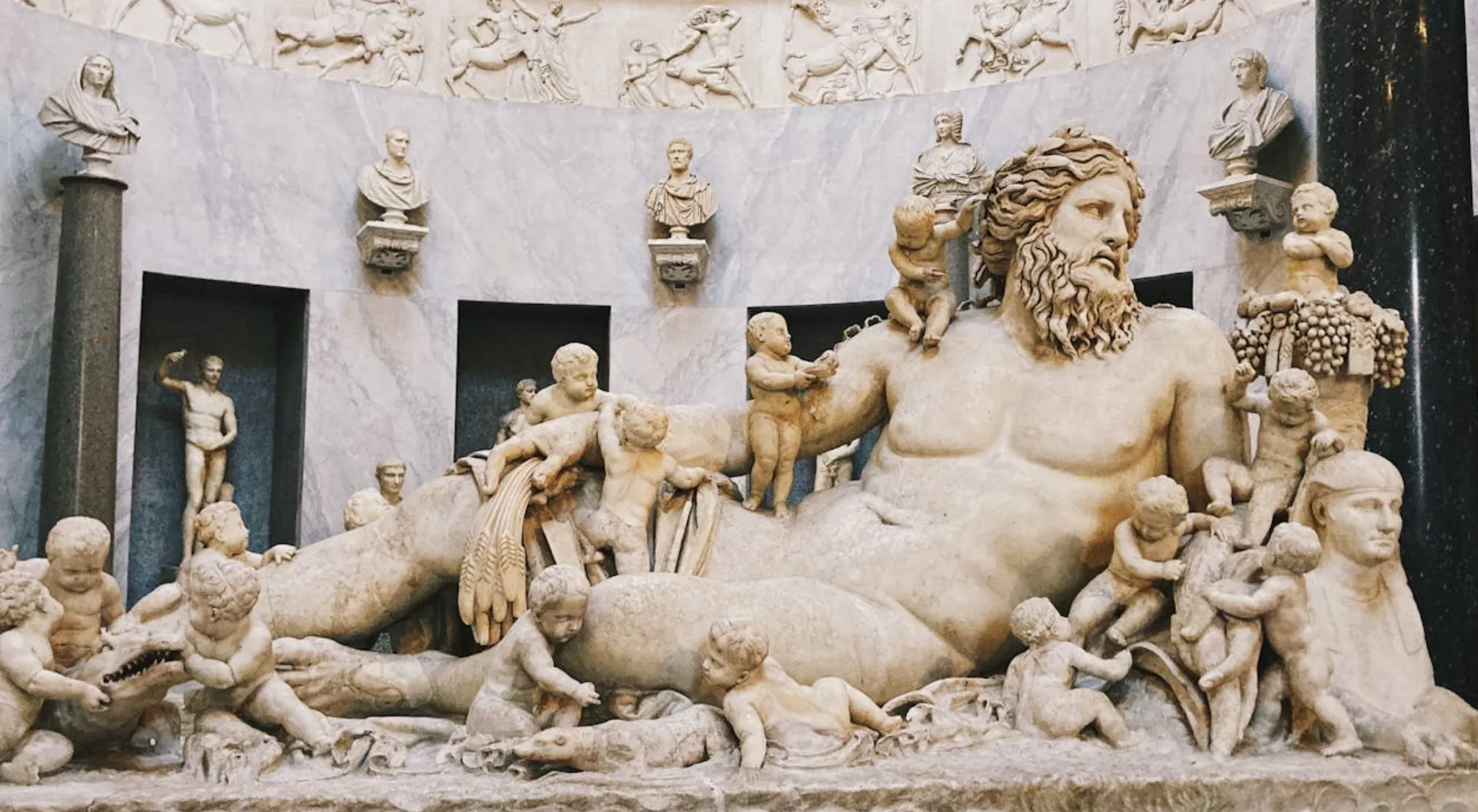 Ancient Roman gods sculpture, a large bearded deity reclines with many cherubic figures. World in Numbers.