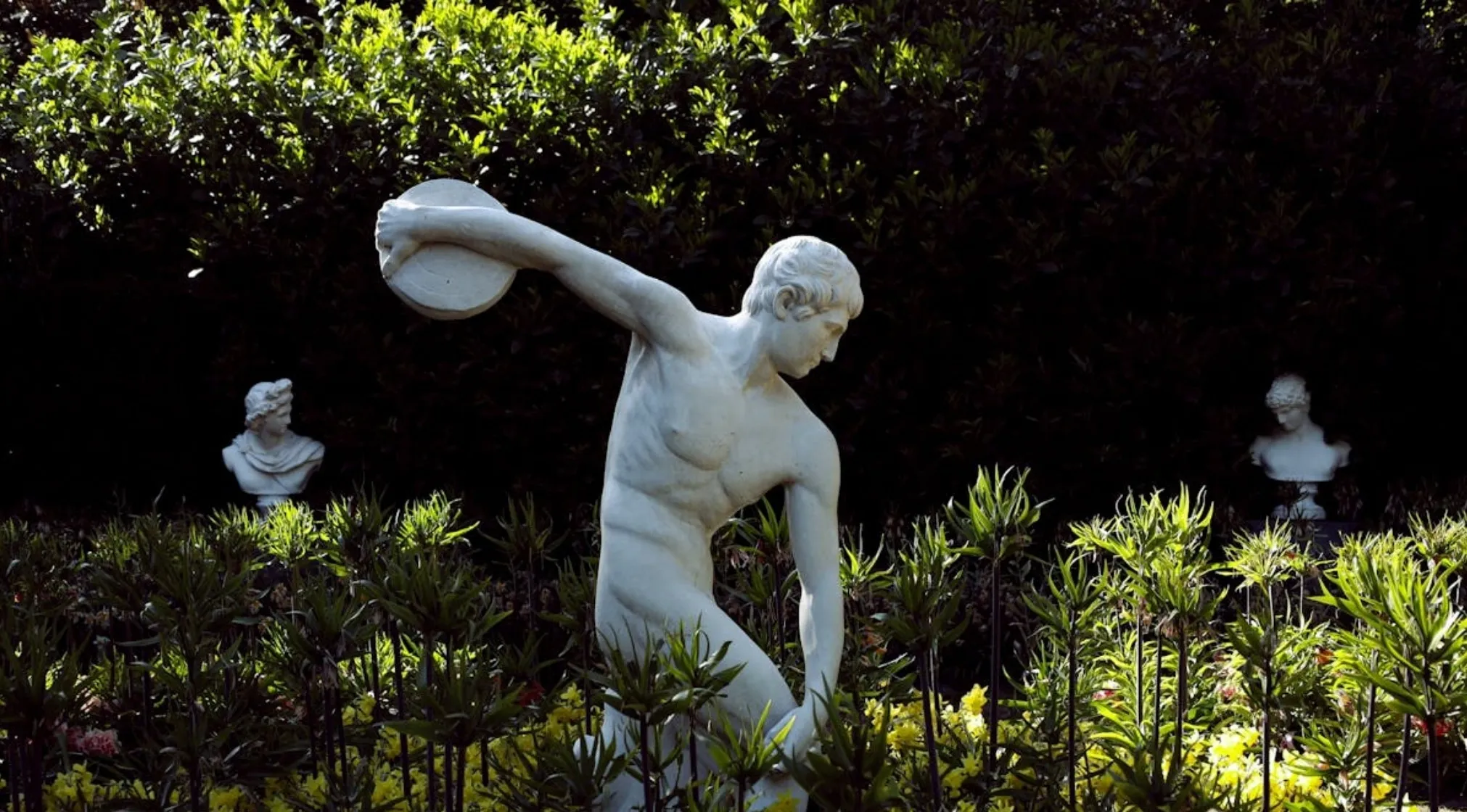 A white statue of a discus thrower in a garden. The olympic symbol and its rings relate to ancient games.