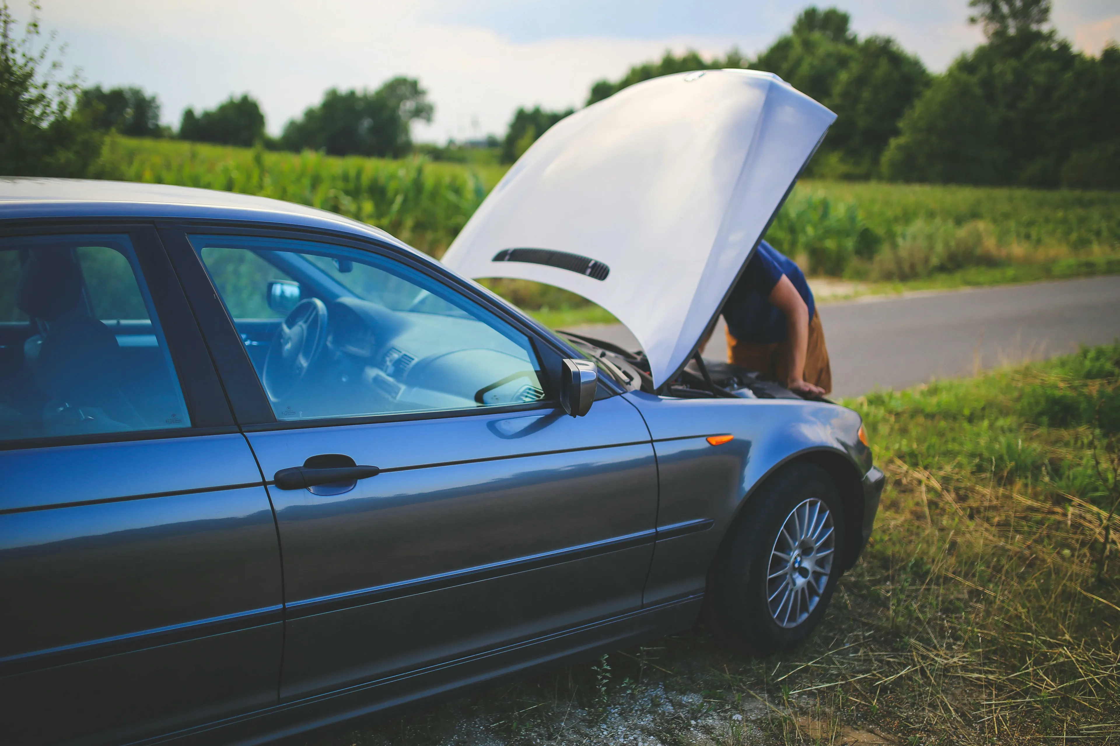 A person looks under the raised hood of a car with a broken down engine, stuck on a rural road. Medieval Profession.