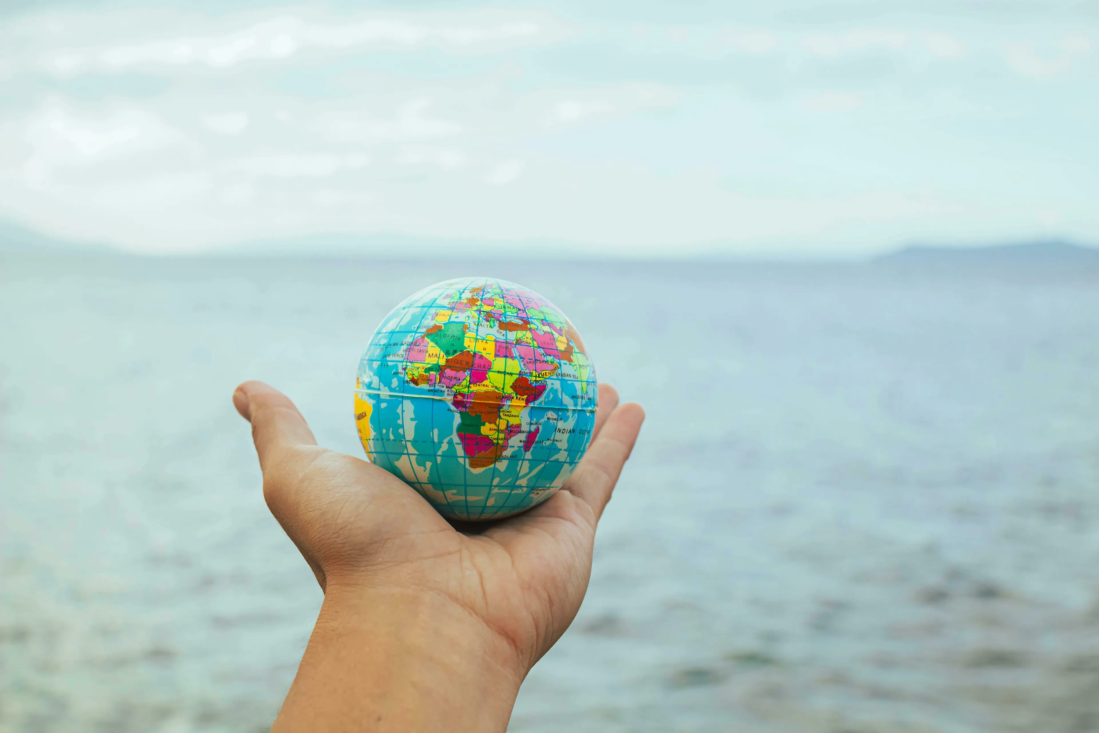 A hand holding a small, colorful globe against a serene sea and sky backdrop. Reflecting on medieval profession and the world.
