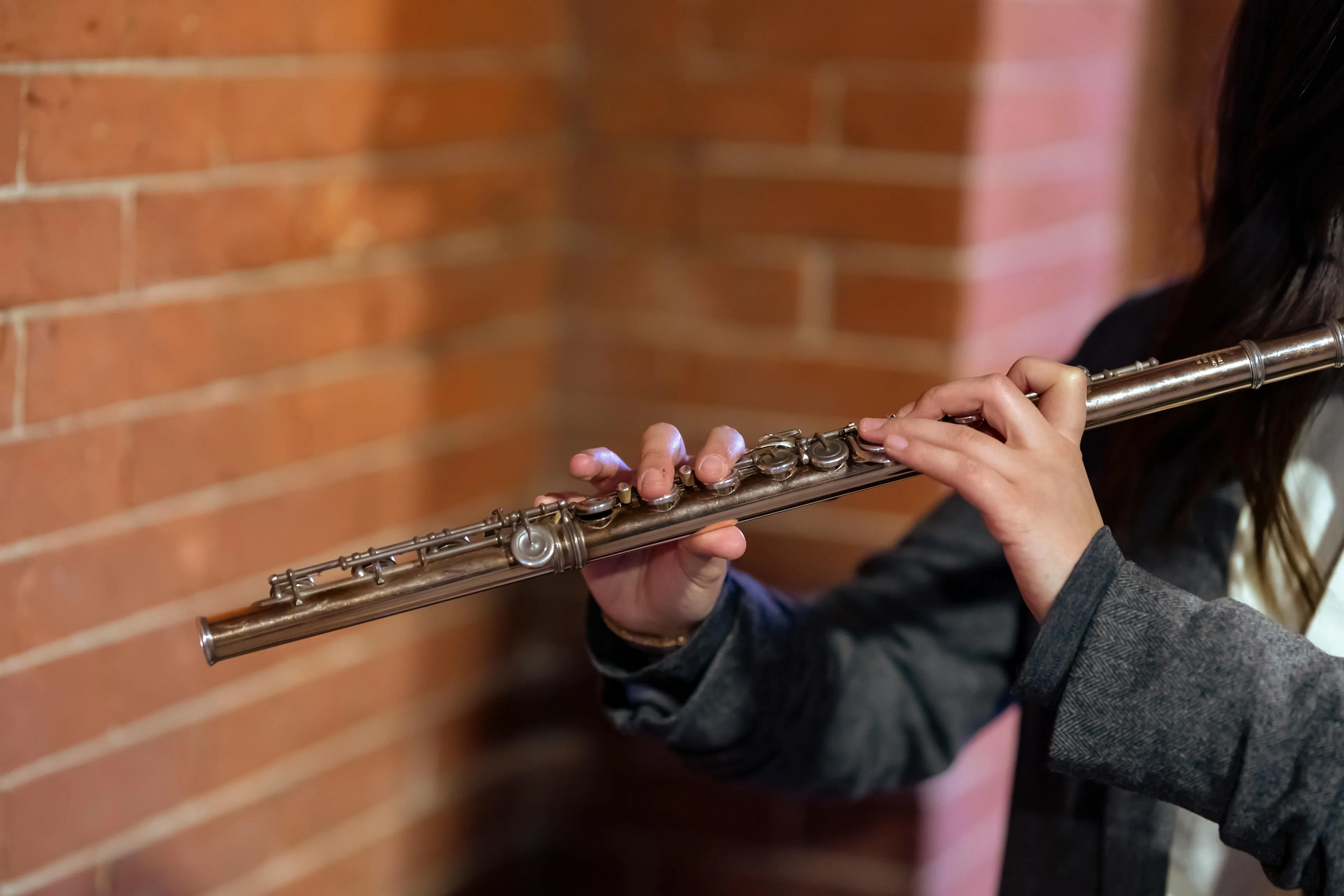 A person's hands holding and playing a silver flute against a brick wall background. Playing an instrument.