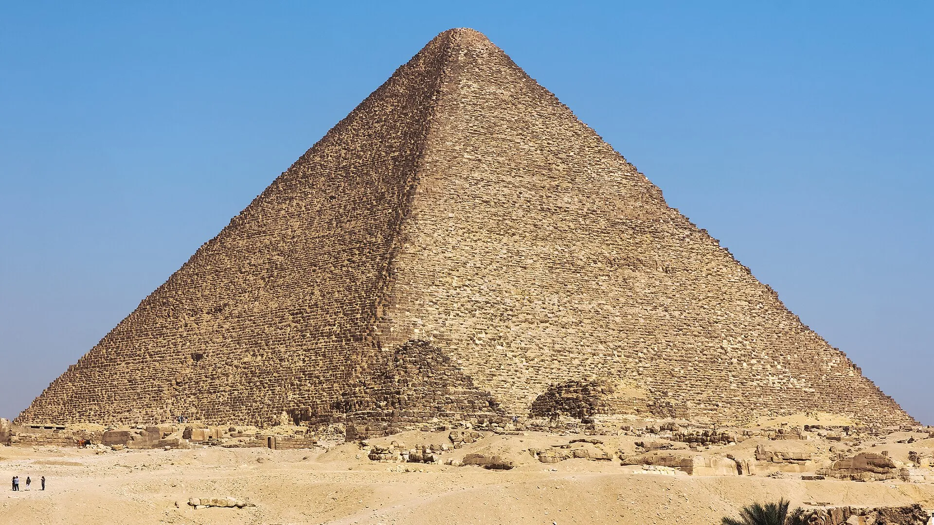 The majestic Great Pyramid of Giza under a clear blue sky, rising from the desert landscape with small figures at its base.
