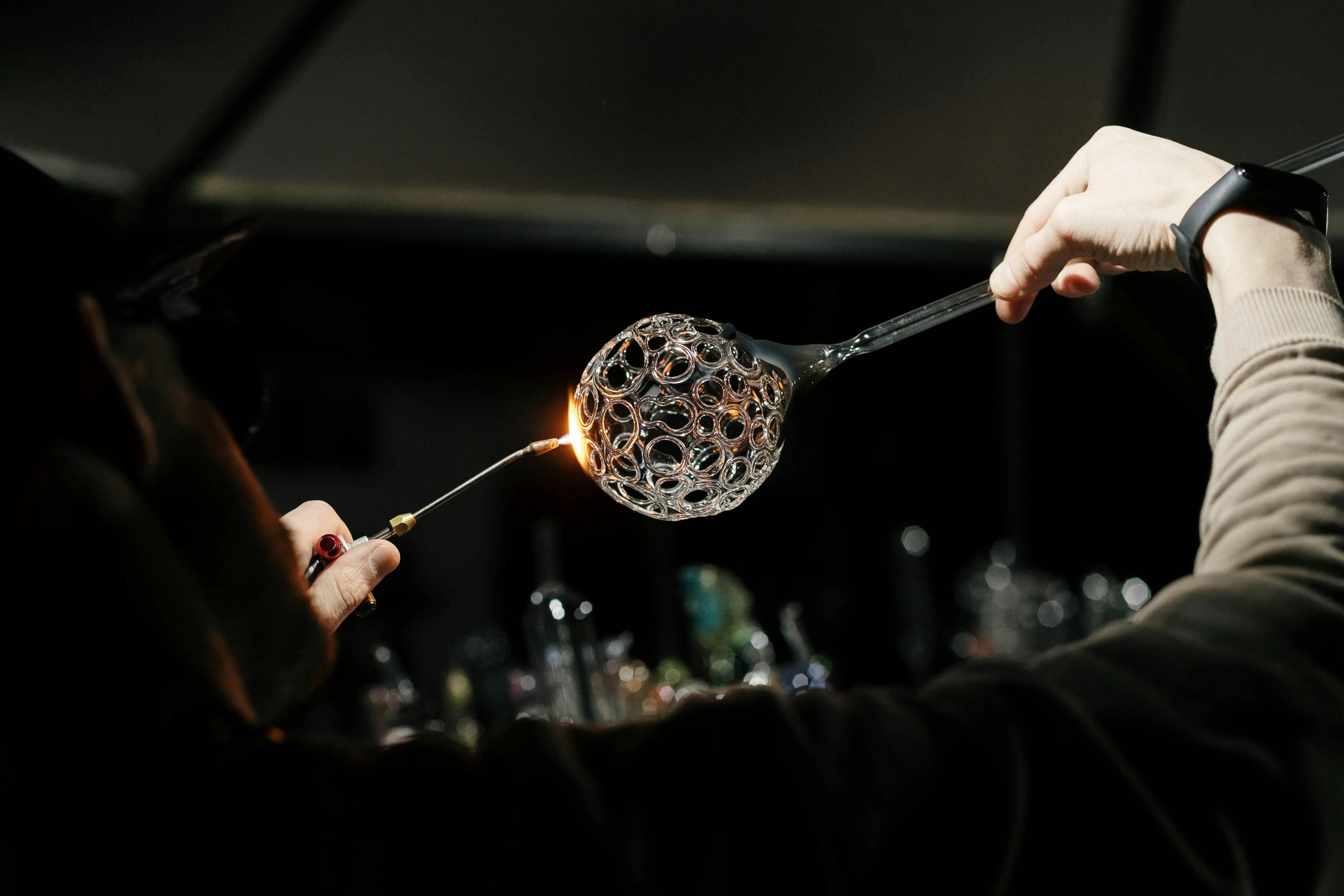 A glassblower skillfully shaping a clear, intricate glass sphere with small holes using a torch in a dark setting.