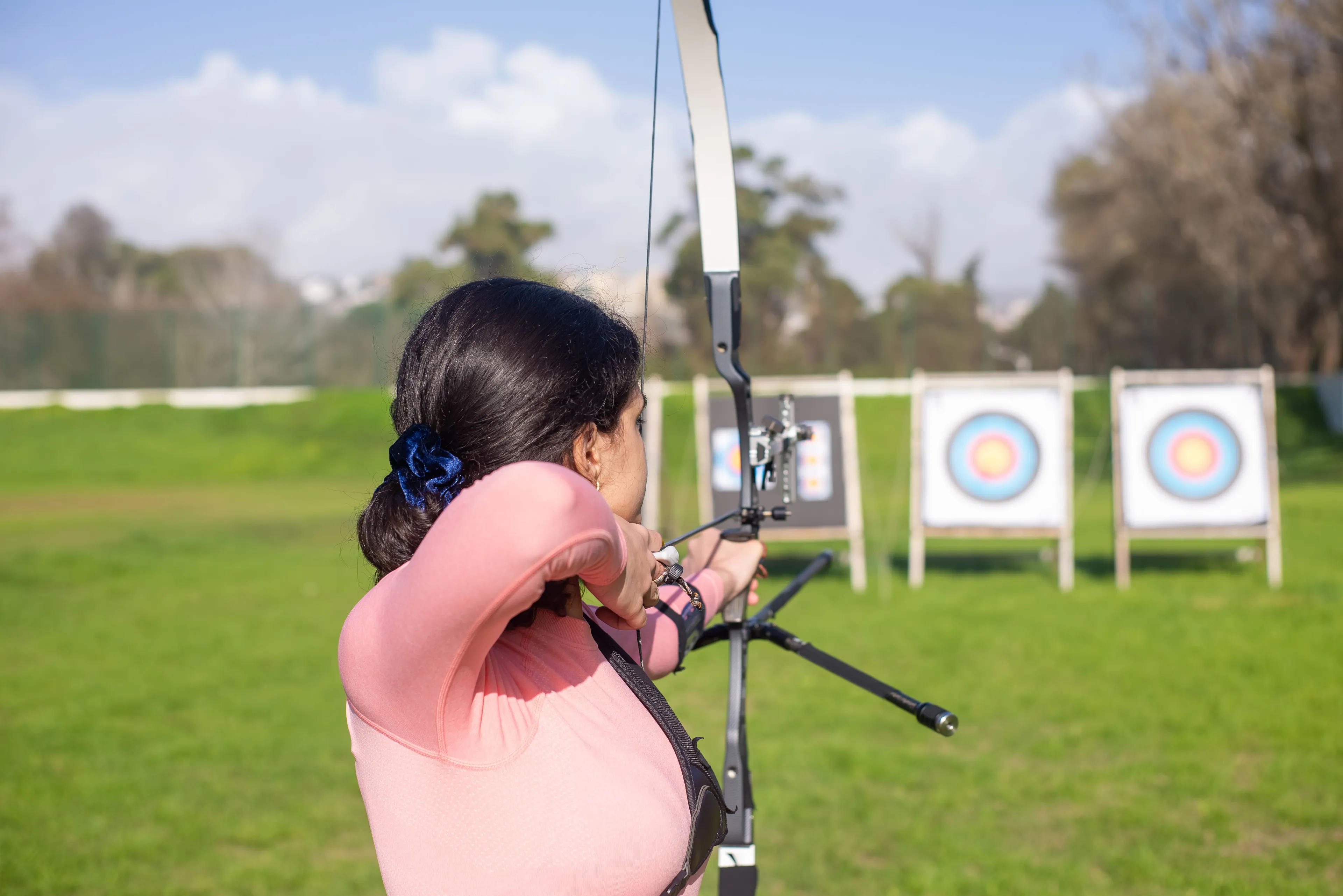 Woman in a pink shirt practices archery, aiming a bow at targets on a sunny green field.
