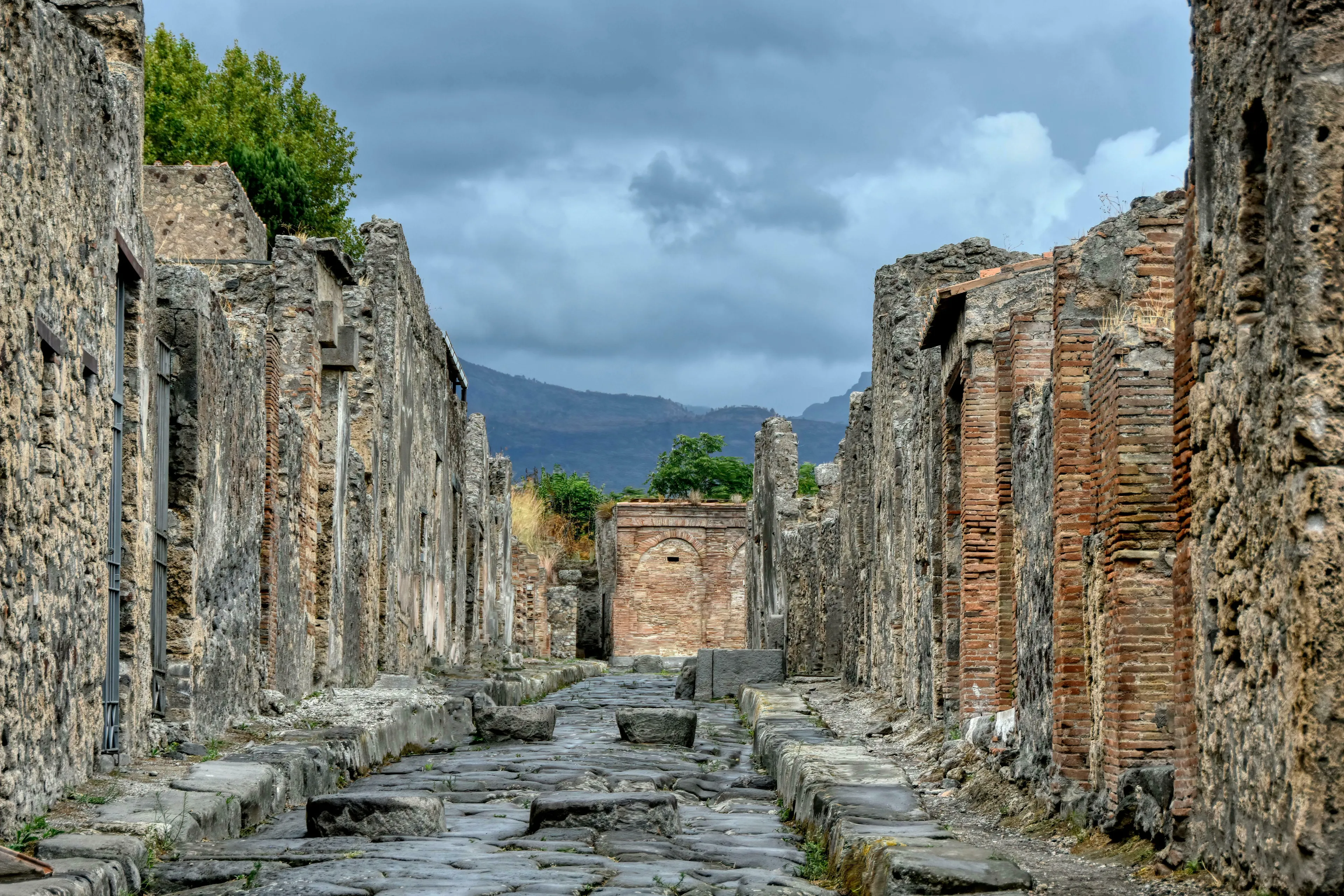 Ruins of Pompeii, an ancient street with stone walls and cobblestones under a cloudy sky. General knowledge quiz hint.