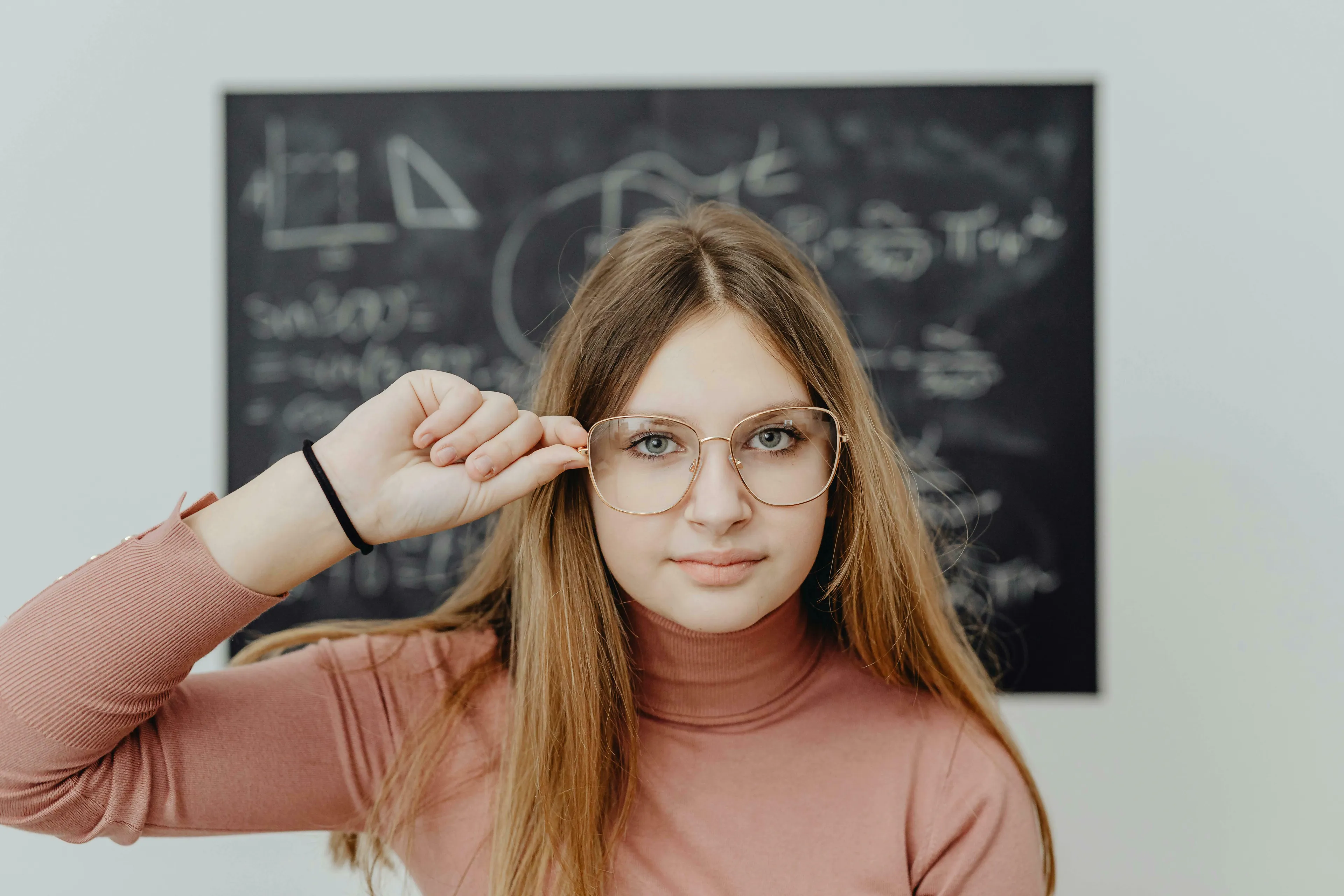 A young woman in a pink turtleneck and gold-rimmed glasses, looking thoughtfully at the camera. Chalkboard with math visible in background. General Knowledge Quiz.