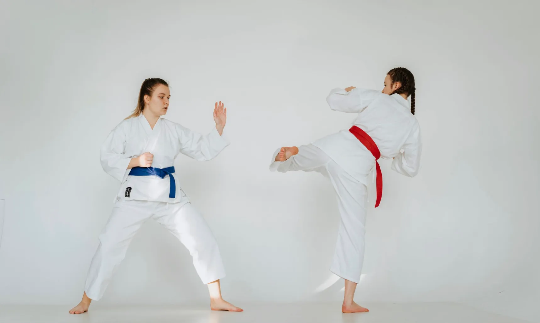 Two women practicing Karate, one with a blue belt blocking a kick from the other with a red belt, showcasing the martial art.