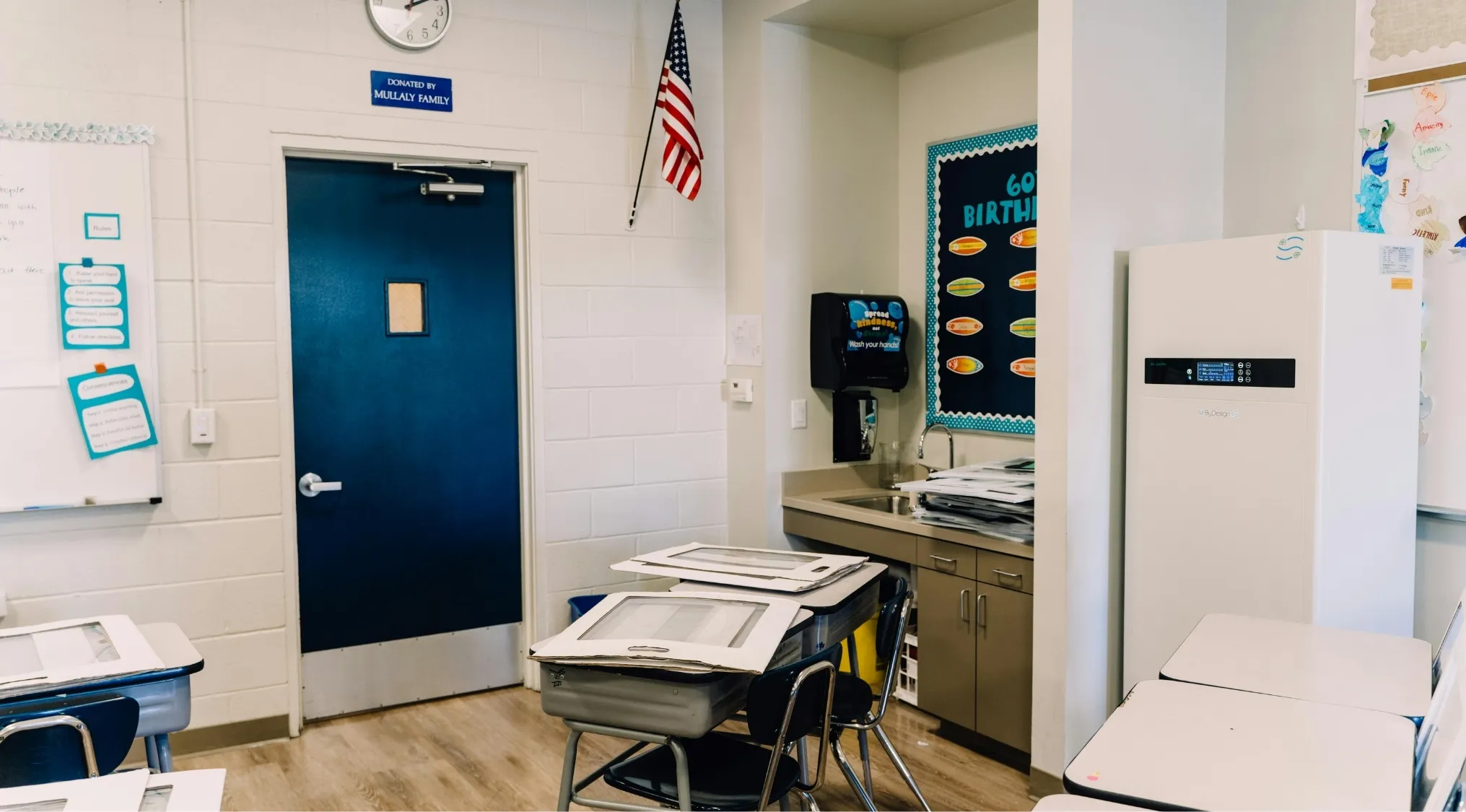 A classroom with an American flag, desks, whiteboard with rules, and a clean air system. Living in America.