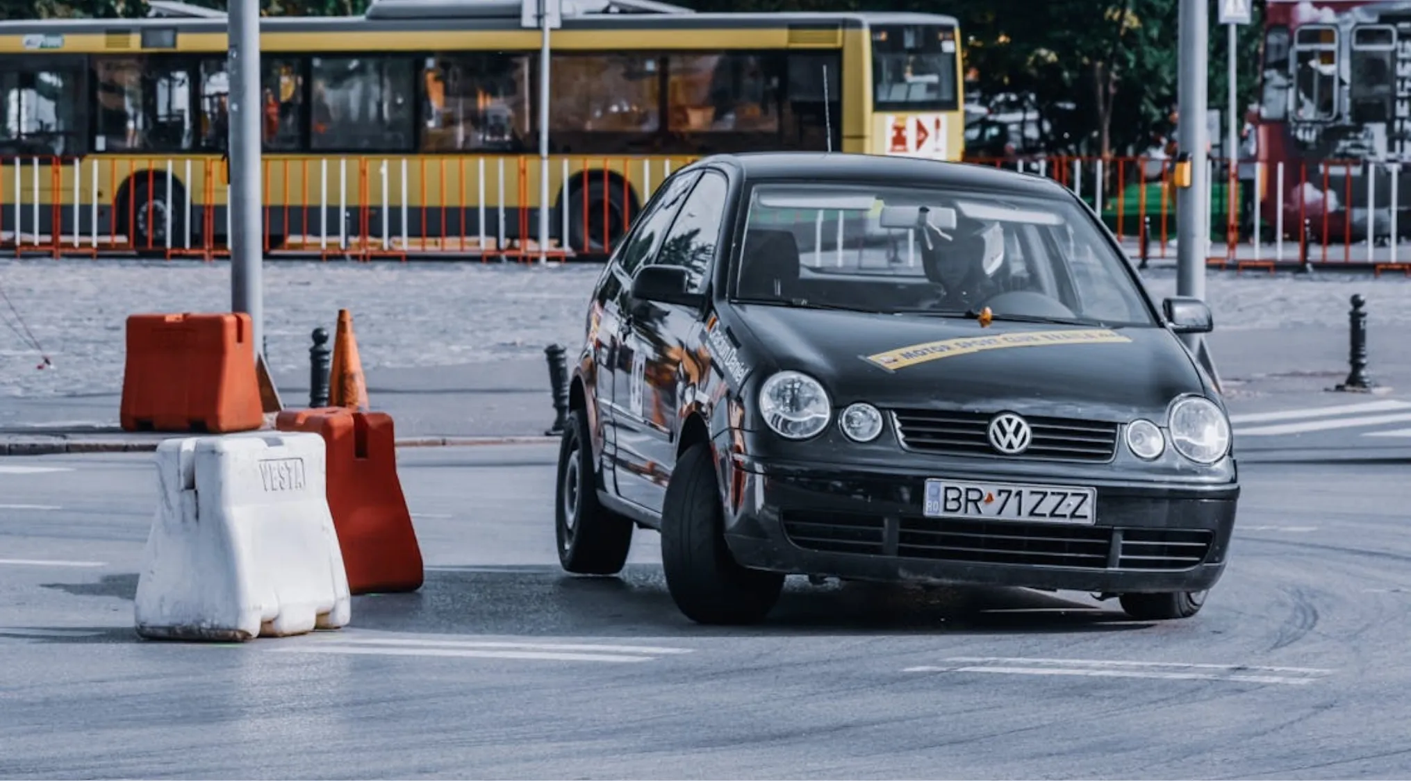 A black Volkswagen driving past orange and white barriers. The image relates to teenagers and provisional driver's licenses.