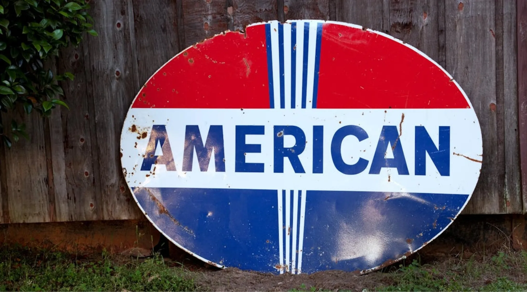 Rustic, distressed American sign, painted red, white, and blue, leaning against a weathered wooden fence. Living in America.