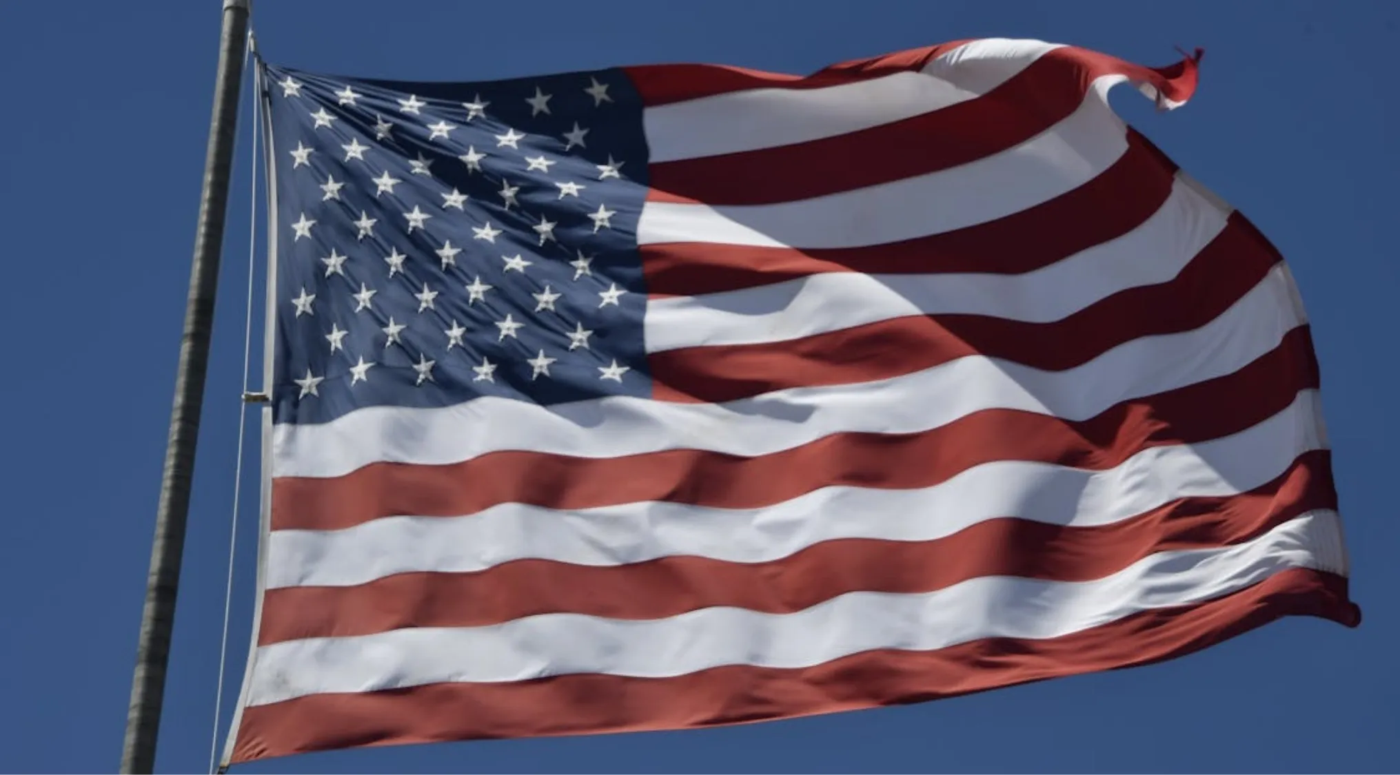 American flag waving against a clear blue sky, highlighting the red and white stripes. Living in America.
