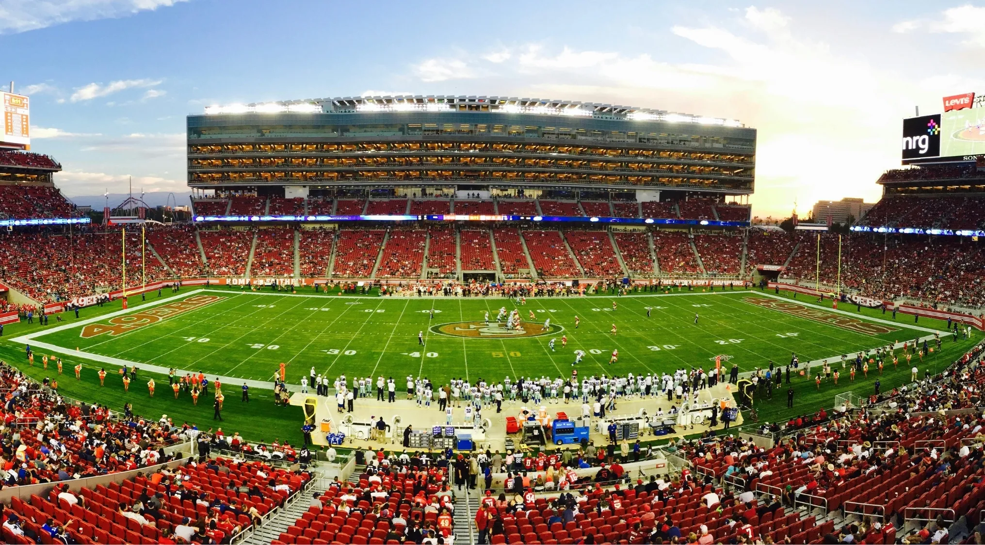 A football field with end zones, packed with fans in a stadium at sunset. Living in America, this highlights American football.