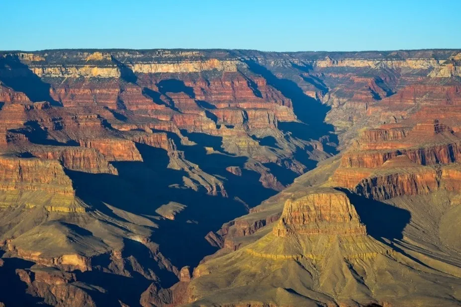 Vast panoramic view of the Grand Canyon, showcasing its layered, colorful rock formations under a clear blue sky.