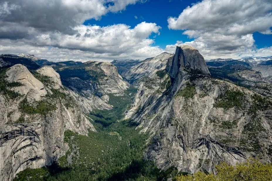 A wide, elevated view of Yosemite National Park with Half Dome visible, showcasing rugged mountains and a lush valley under a partly cloudy sky.