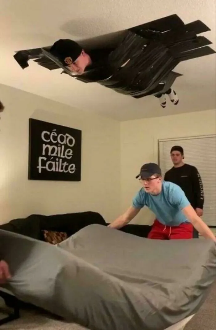 Person taped to ceiling with black duct tape while others prepare a bed