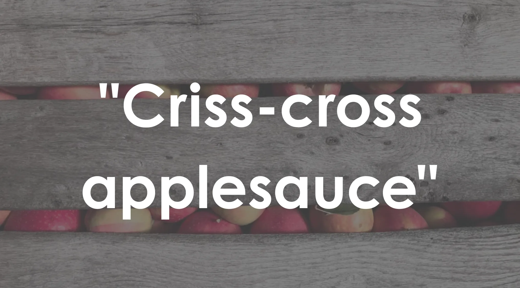 White text "Criss-cross applesauce" over a blurred wooden background with hints of red and yellow. Living in America.