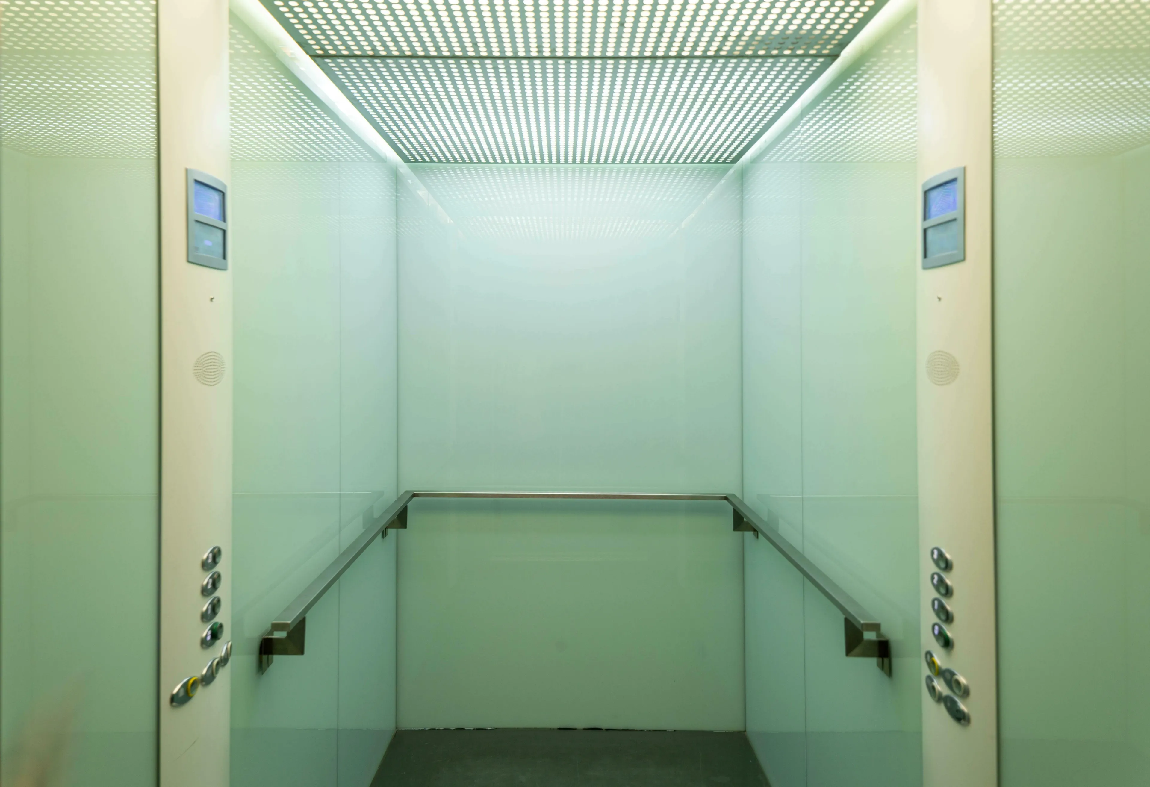 A modern, empty elevator with green-tinted glass walls and a patterned ceiling, useful for American vs British English words quiz.