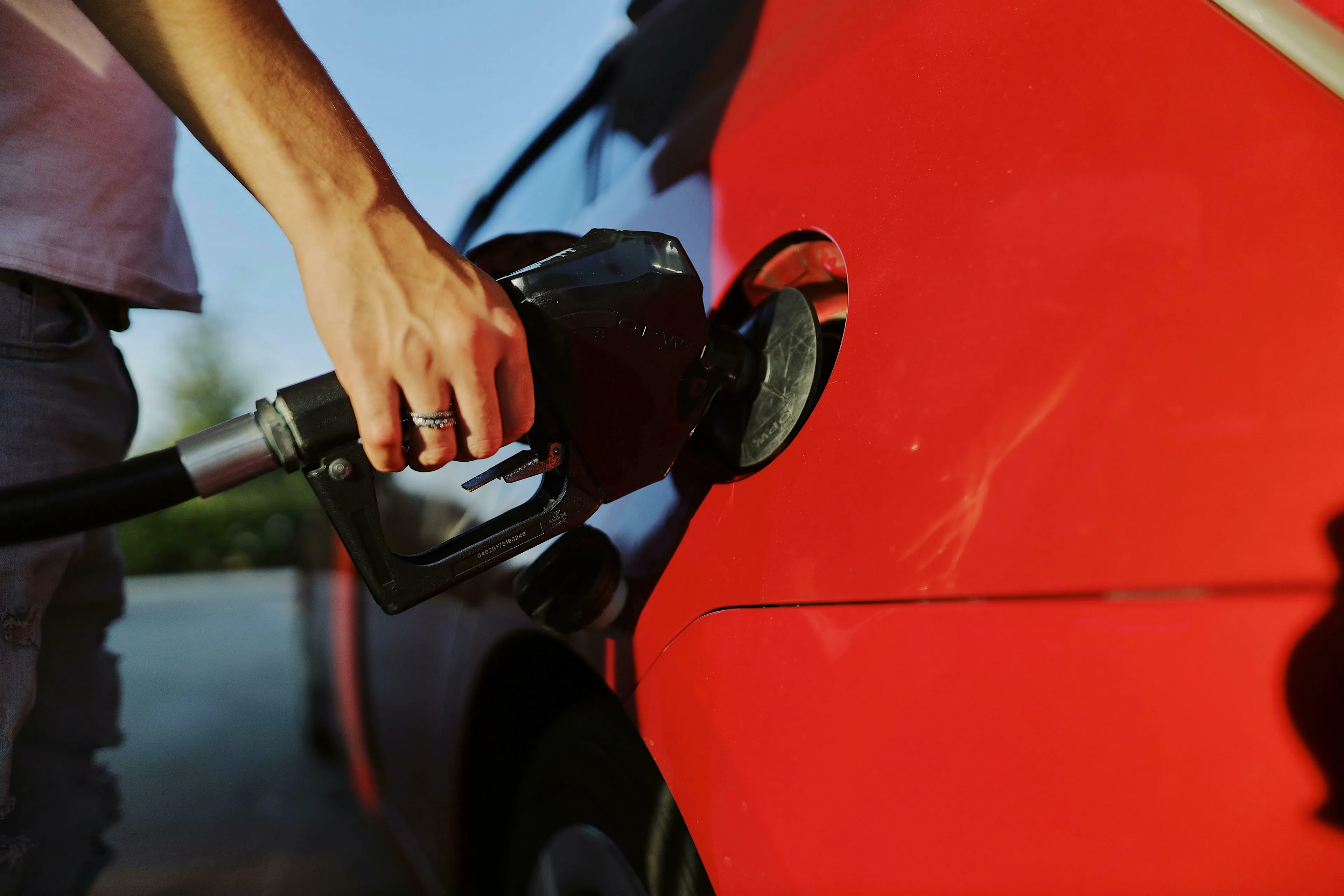 Close-up of a person's hand with a ring, holding a black fuel nozzle and filling a red car. American vs British English words quiz.