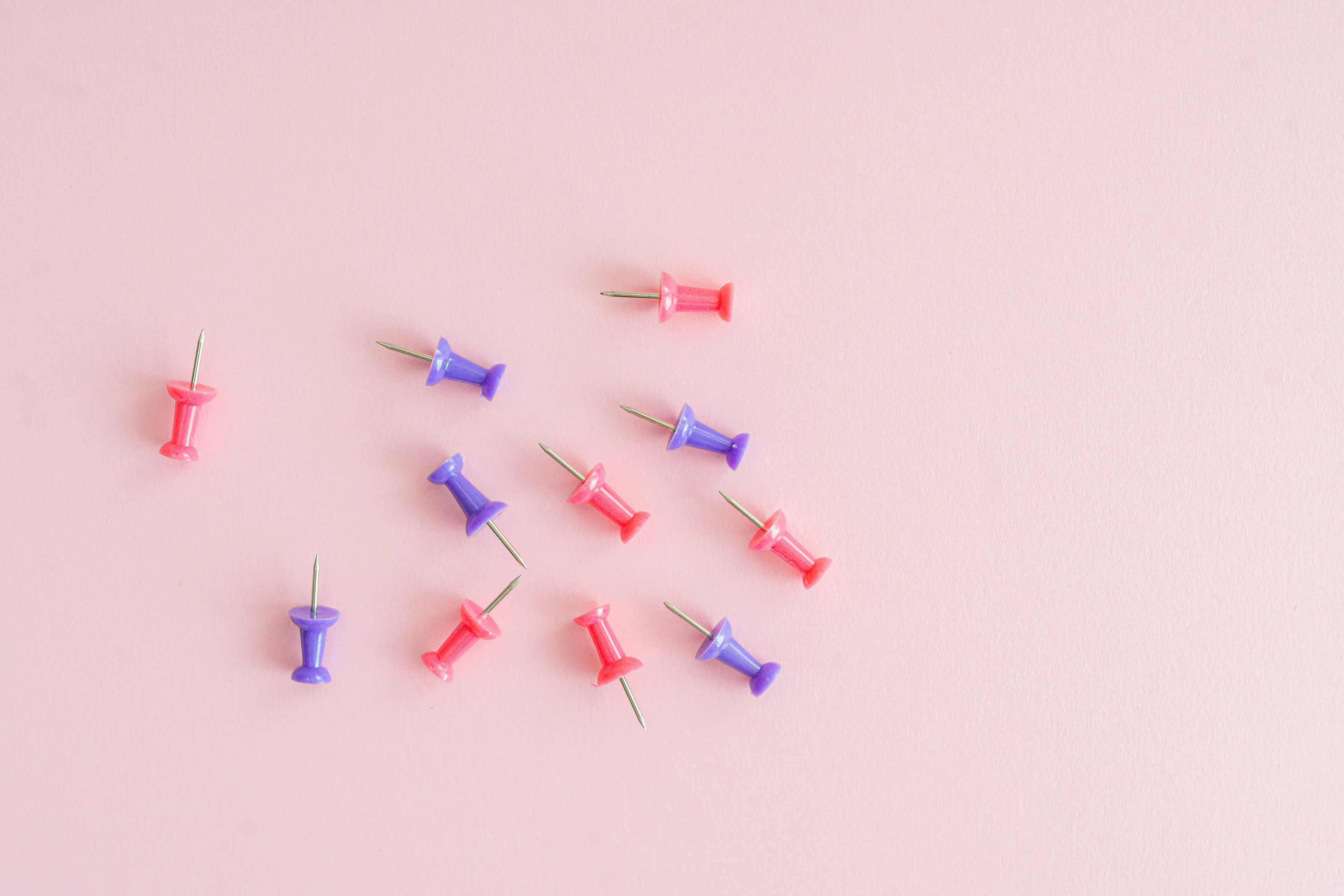 Pink and purple thumbtacks scattered on a light pink background, perfect for an American Vs British English stationery quiz.