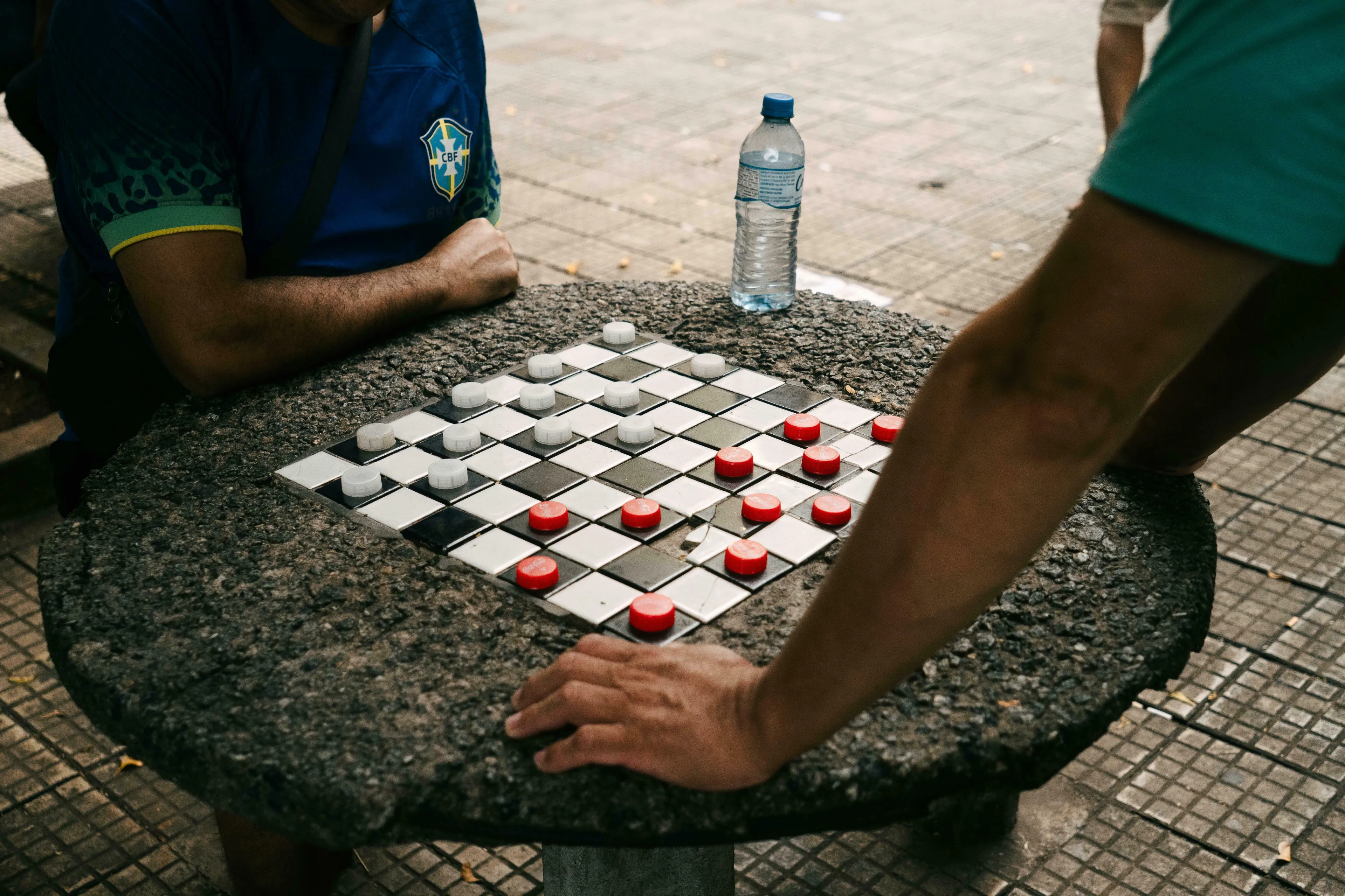 Two men playing checkers, or draughts, on a stone table with red and white pieces, a water bottle nearby.
