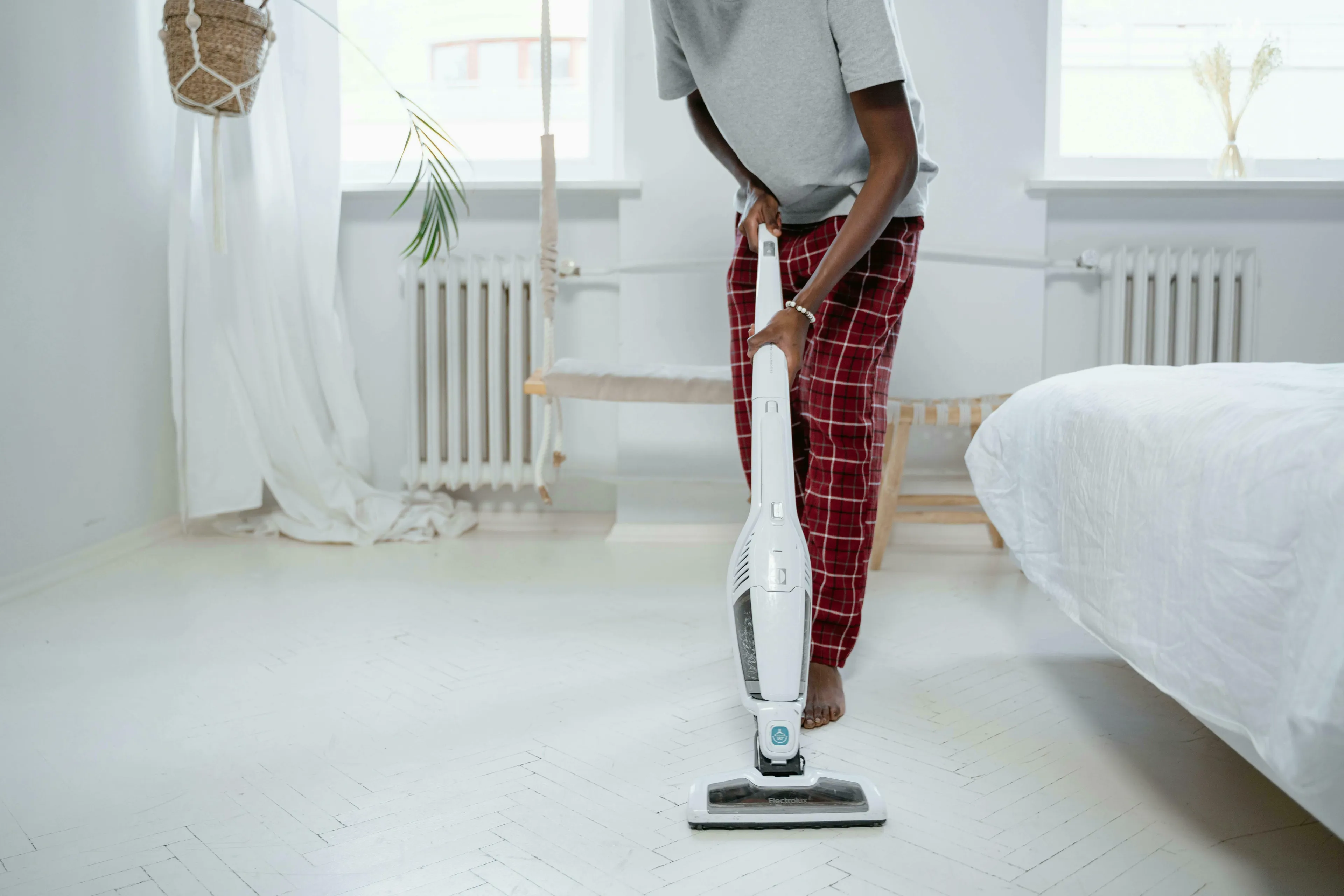 A person in a gray shirt and red plaid pants uses a white stick vacuum on a white floor. American Vs British English Words Quiz.