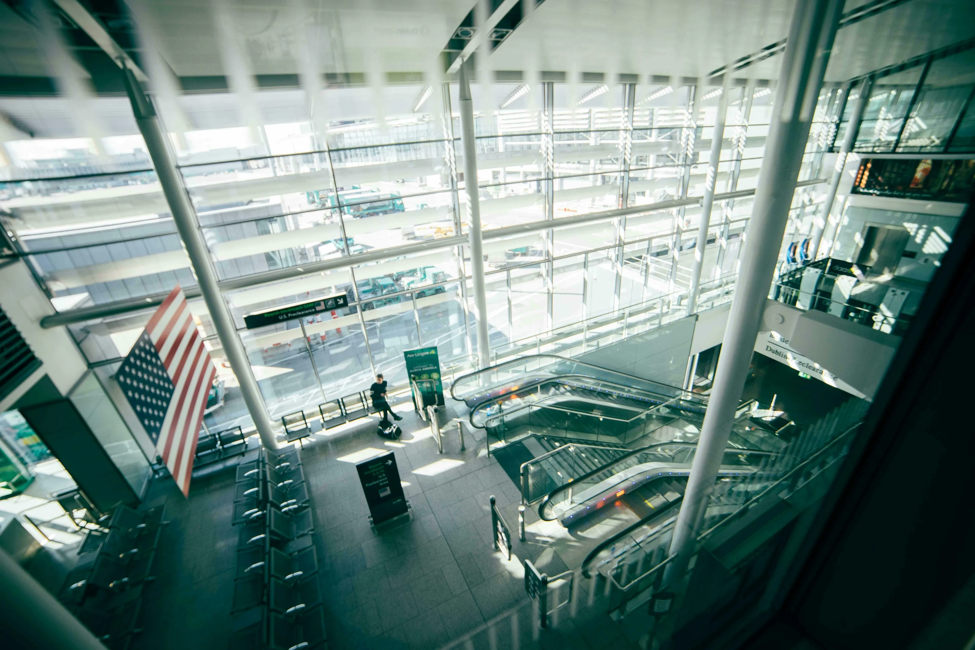 Modern American airport terminal with a US flag, escalators, and a lone traveler with their luggage. American vs British English words quiz.