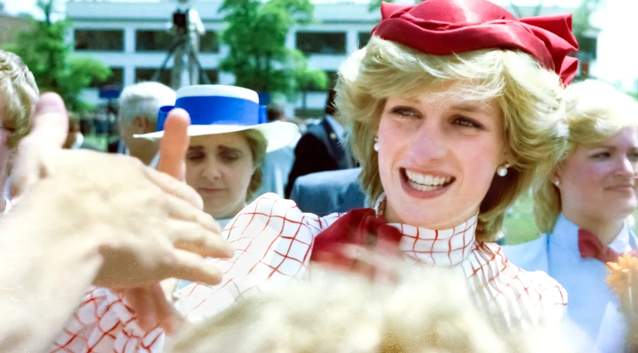 Princess Diana smiling and reaching out to greet people. She wears a red bow hat and a white checkered top.