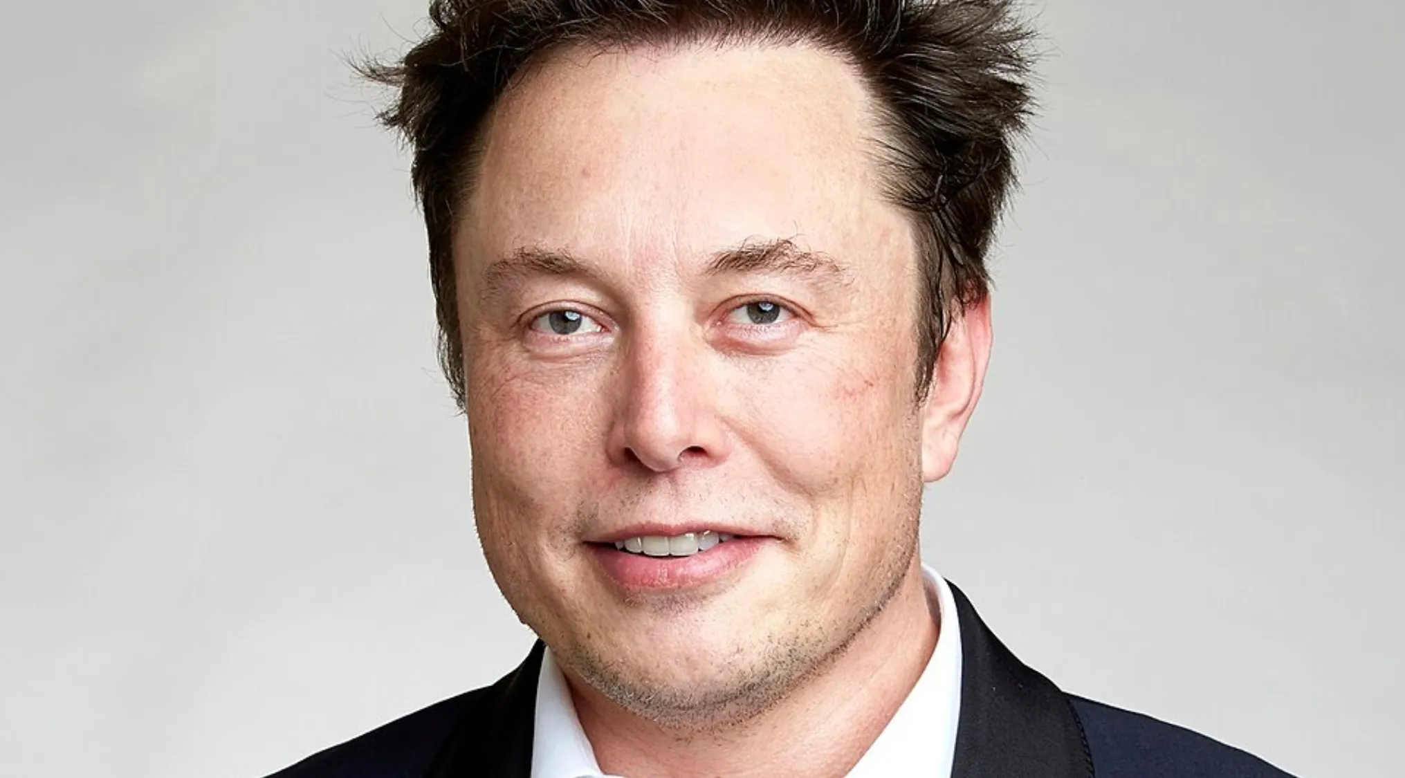 Close-up of Elon Musk, a famous billionaire, in a black suit and white shirt, smiling slightly.