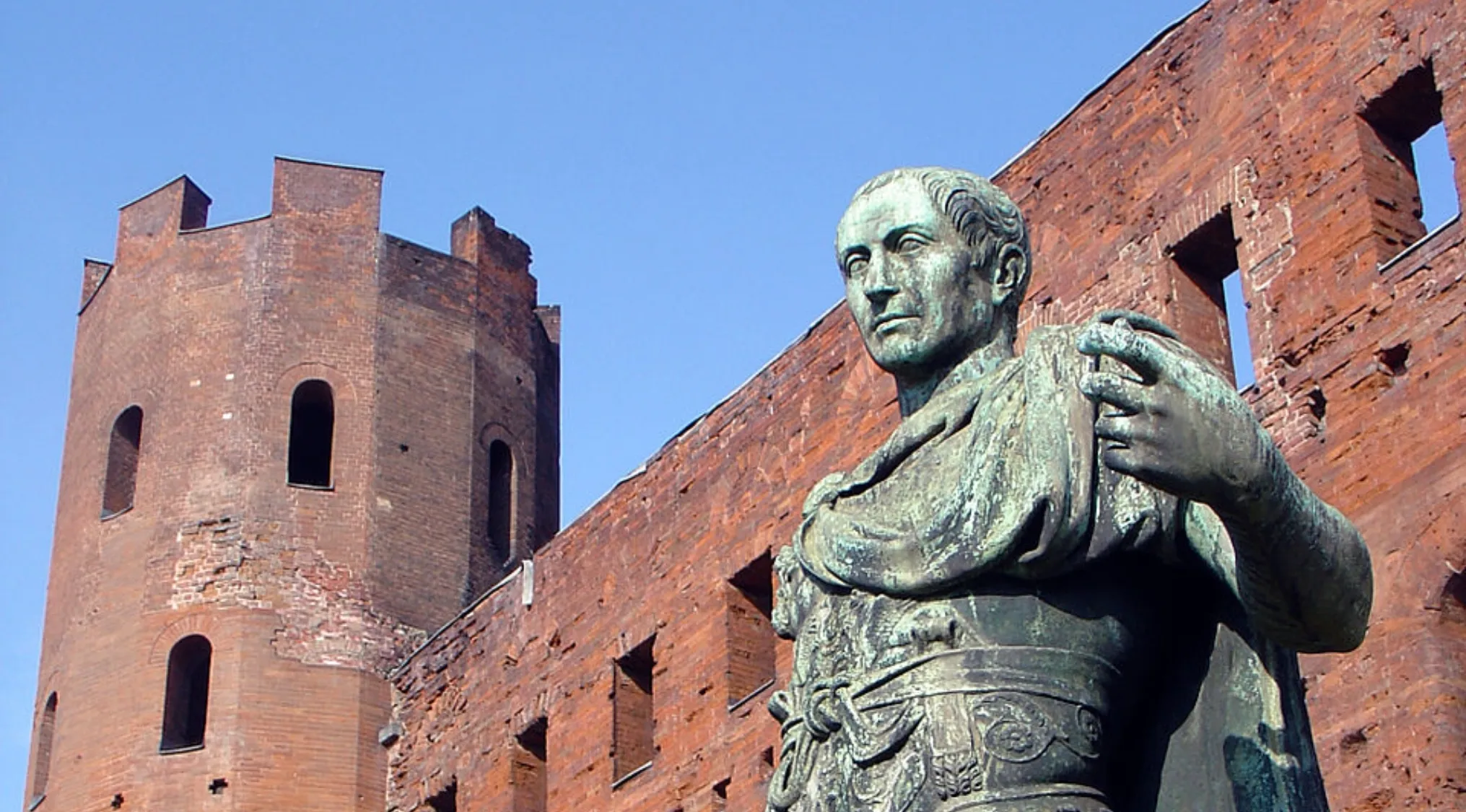 Bronze statue of a Roman general against a clear blue sky, with a red brick ruin in the background.