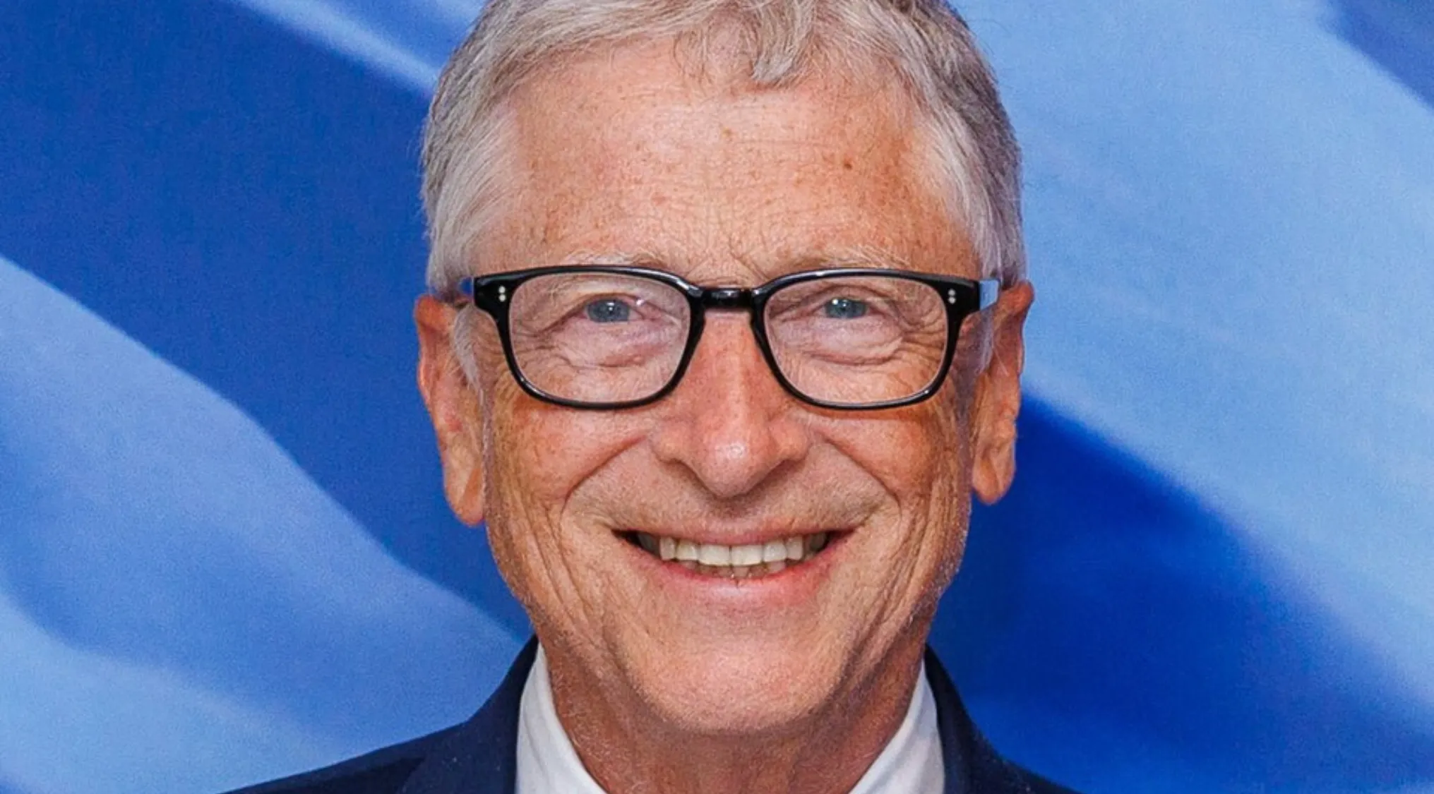 Famous person, Microsoft co-founder, Bill Gates smiling, wearing glasses against a blue background.