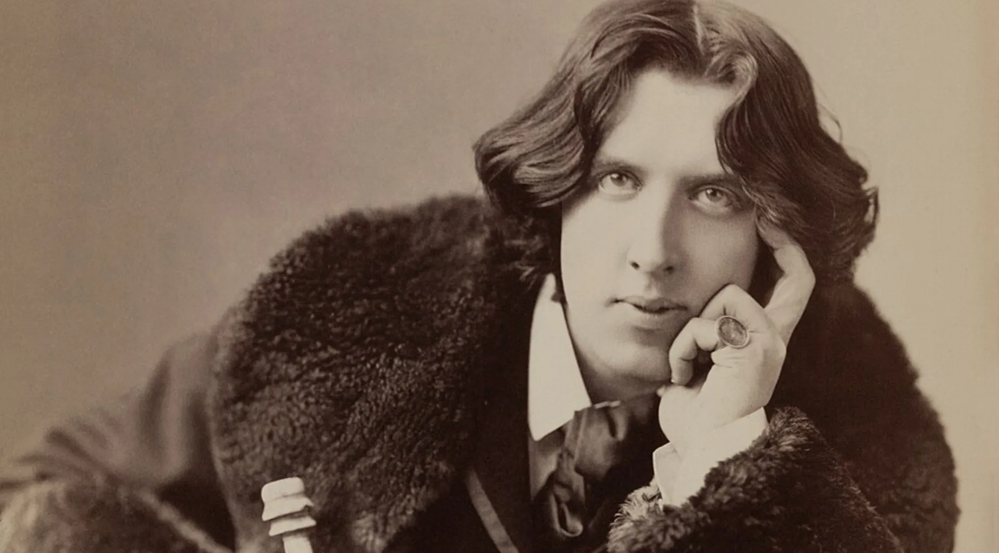 Sepia portrait of a young Oscar Wilde, an Irish writer, with long dark hair, wearing a fur-collared coat.