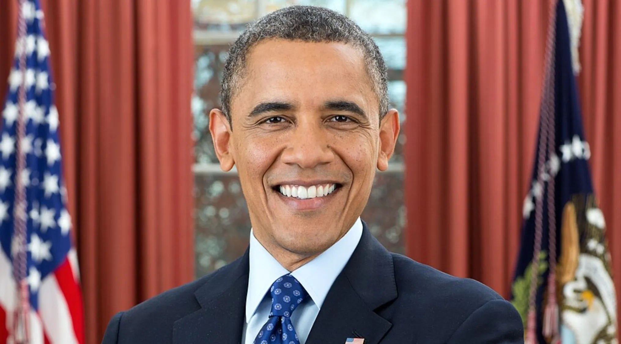 Smiling Barack Obama, the first African American president of the United States, in a suit and tie with flags in the background.