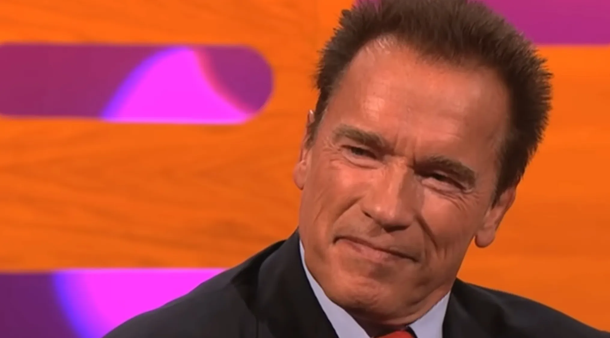 Famous Person Arnold Schwarzenegger, actor and former bodybuilder, smiling in a suit against an orange and purple background.