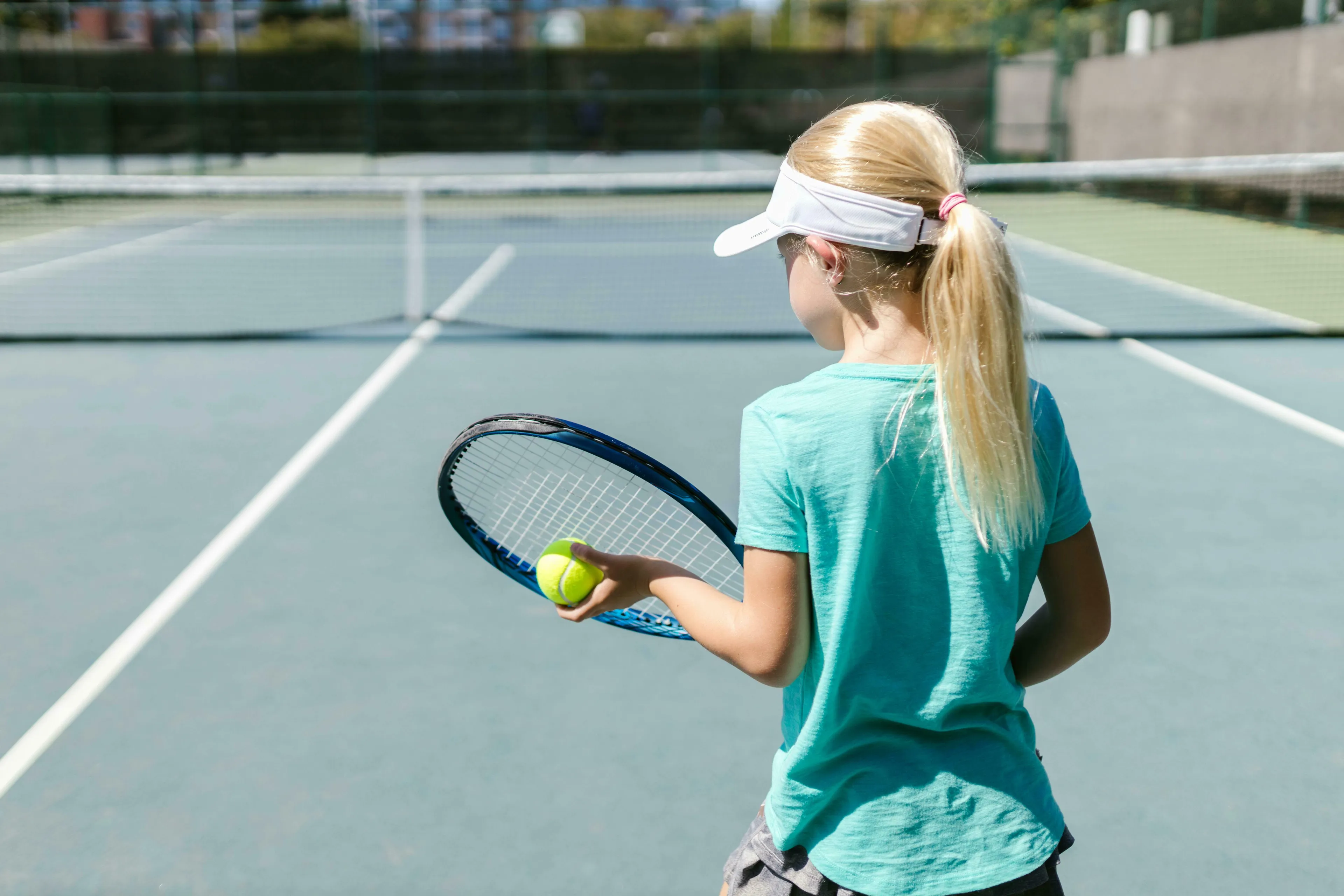 A young girl with blonde hair in a ponytail and a visor, holding a tennis racket and ball on a sunny tennis court.
