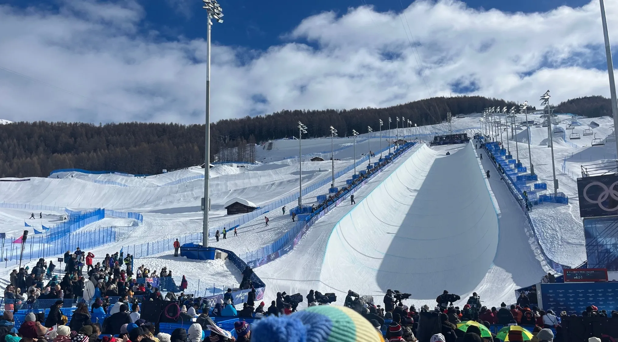 Spectators watch Winter Olympic Games sports event, with a halfpipe and ski slopes under a cloudy sky.