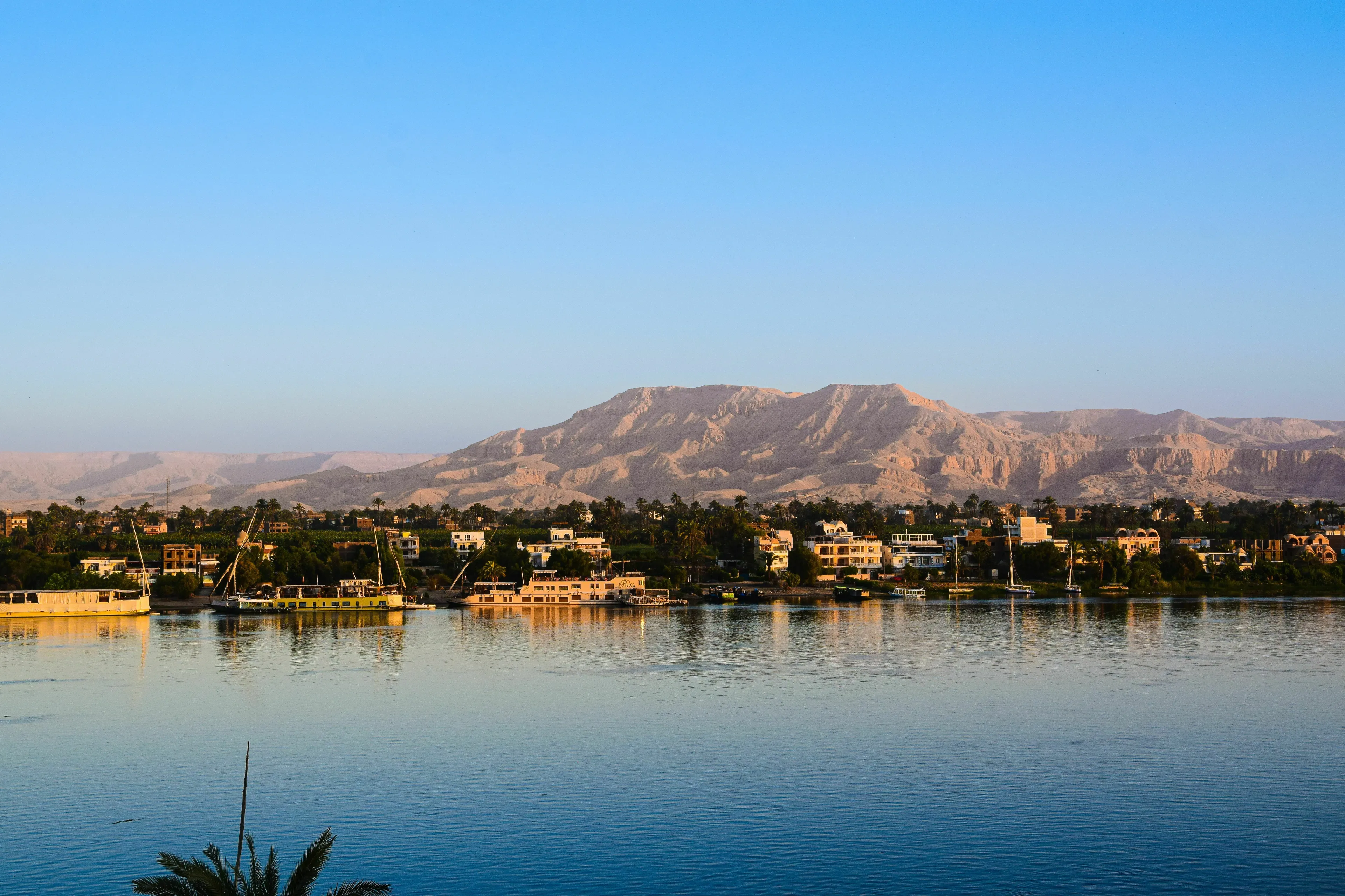 A picturesque view of the Nile River with boats, lush banks, and desert mountains under a clear blue sky. Geography facts.