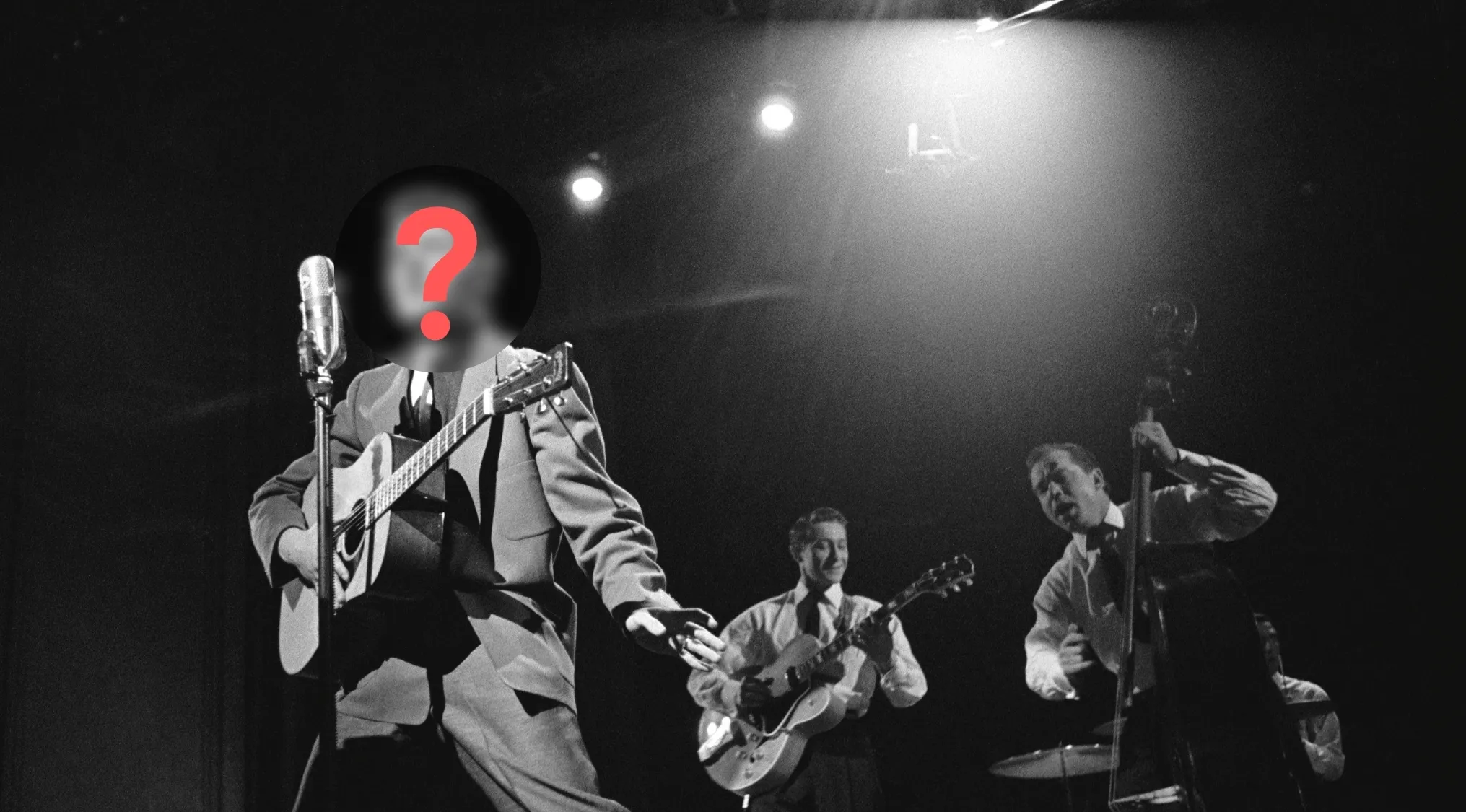 Black and white image of a musician, with a question mark on his face, playing guitar and singing into a microphone. This is Elvis, the King of Rock and Roll.