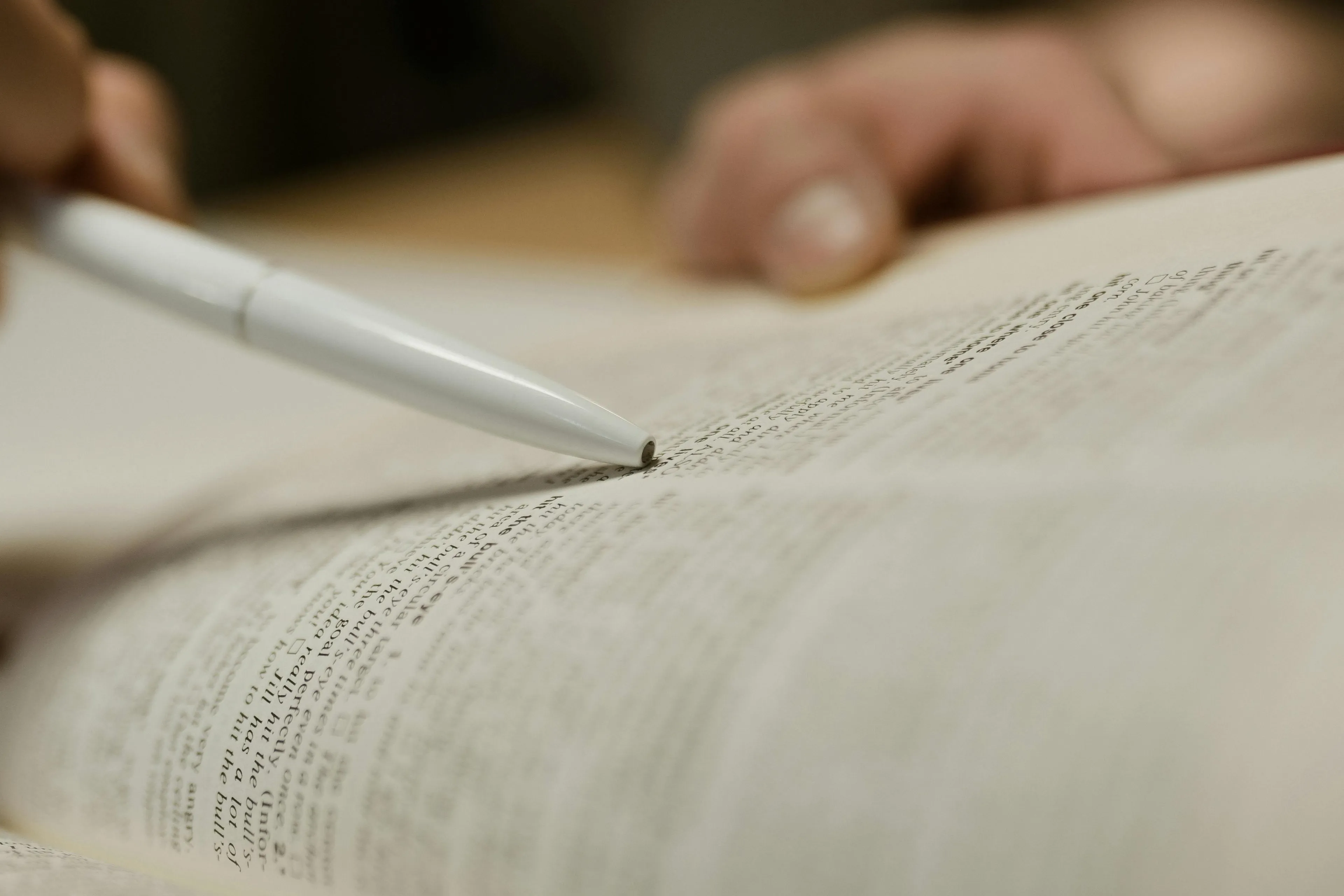 A person uses a white pen to point at text in a book, demonstrating a method for school questions or studying English.