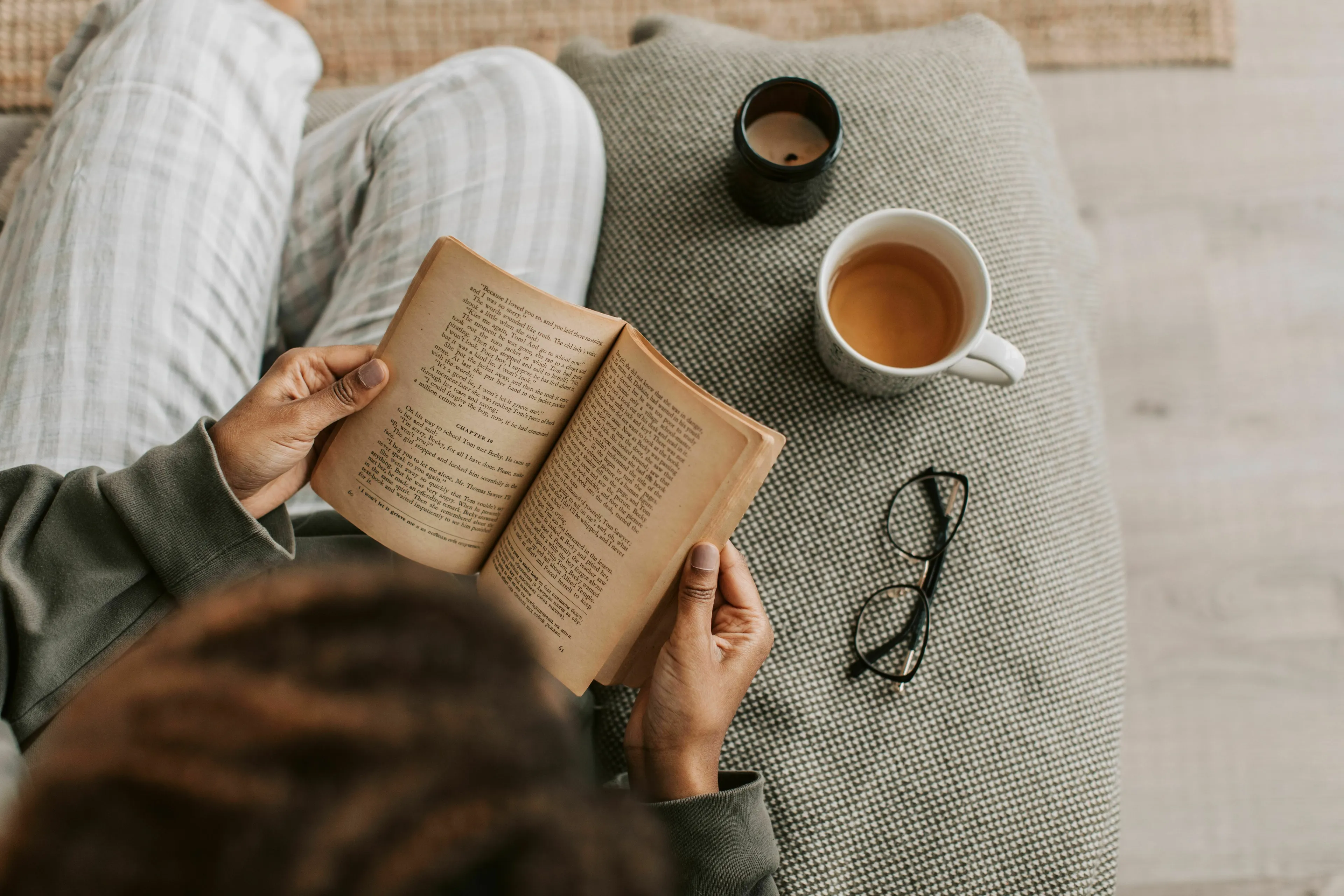 Overhead shot of a person reading an old book on a gray couch, with a teacup, candle, and reading glasses nearby. General knowledge.