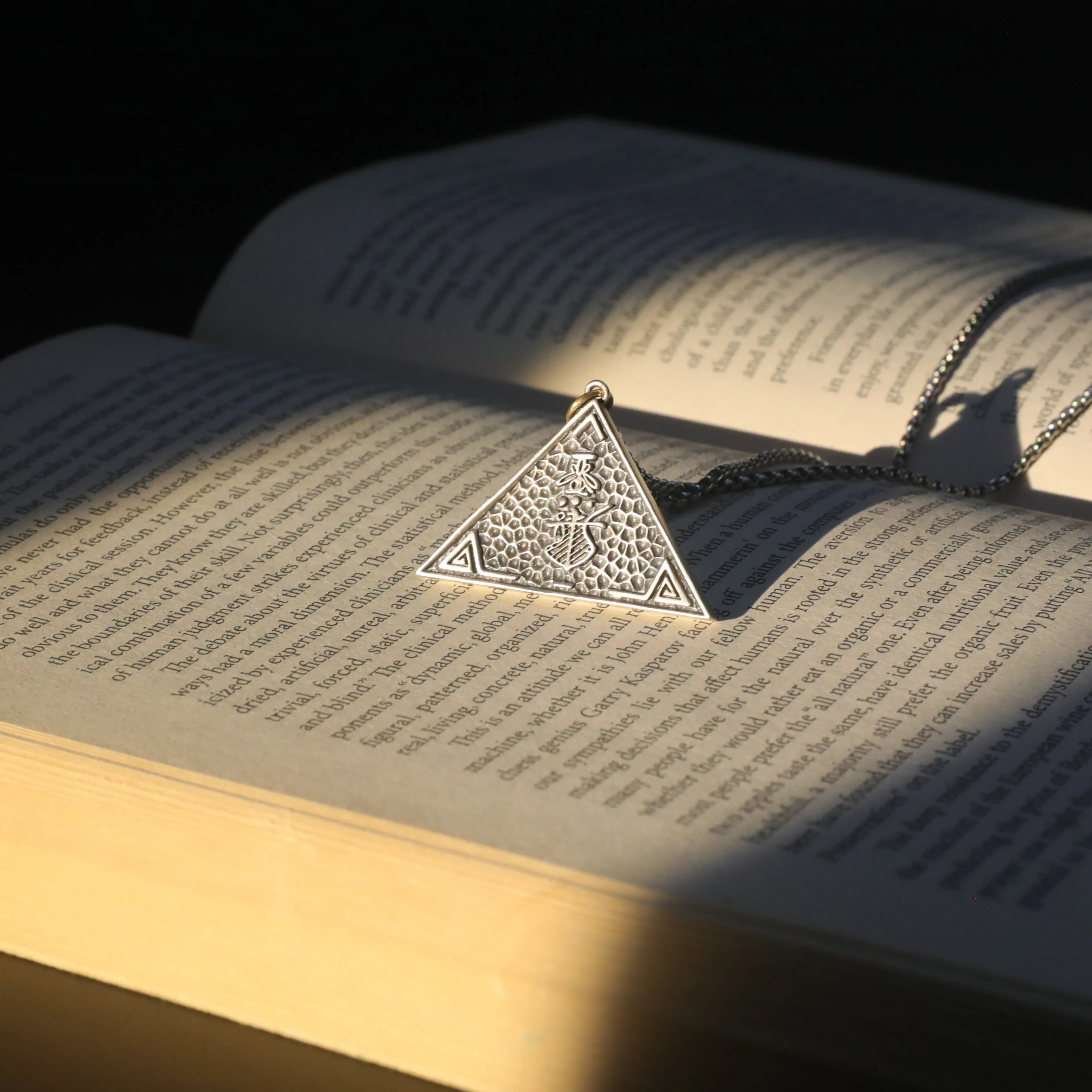A silver, textured pyramid pendant with a symbol, resting on an open book, hinting at general knowledge and secret societies.