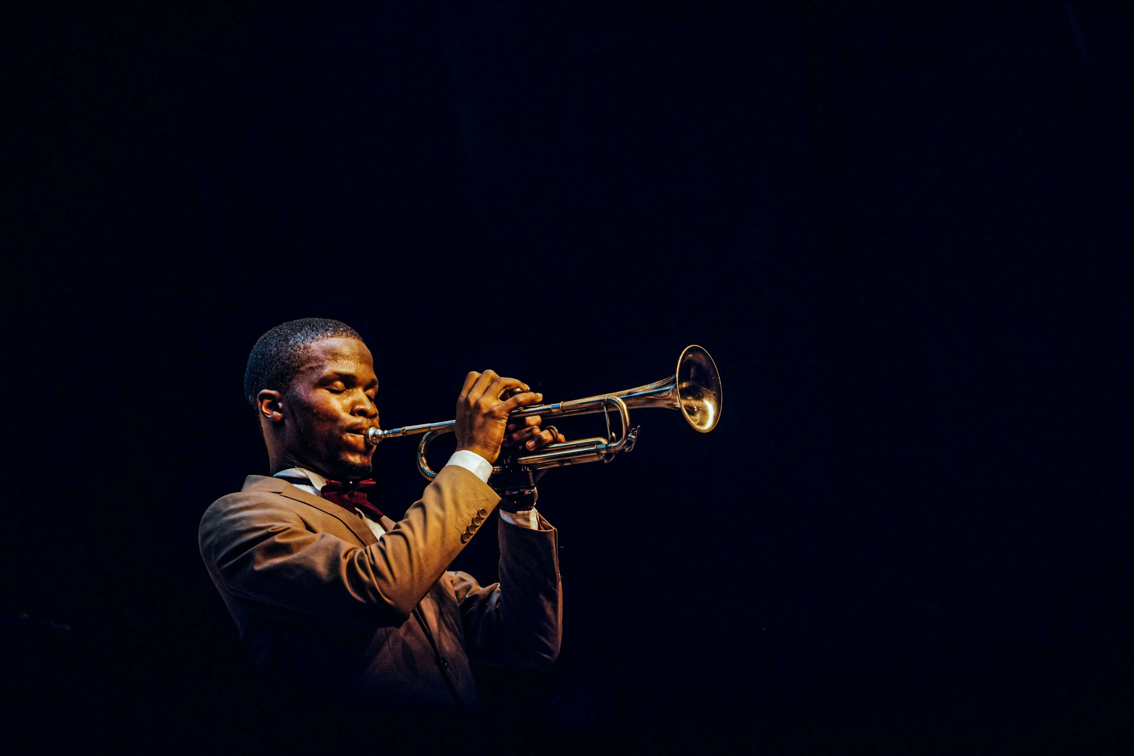 A musician in a suit with a bow tie, playing a trumpet on a dark stage. New Orleans African-American communities.