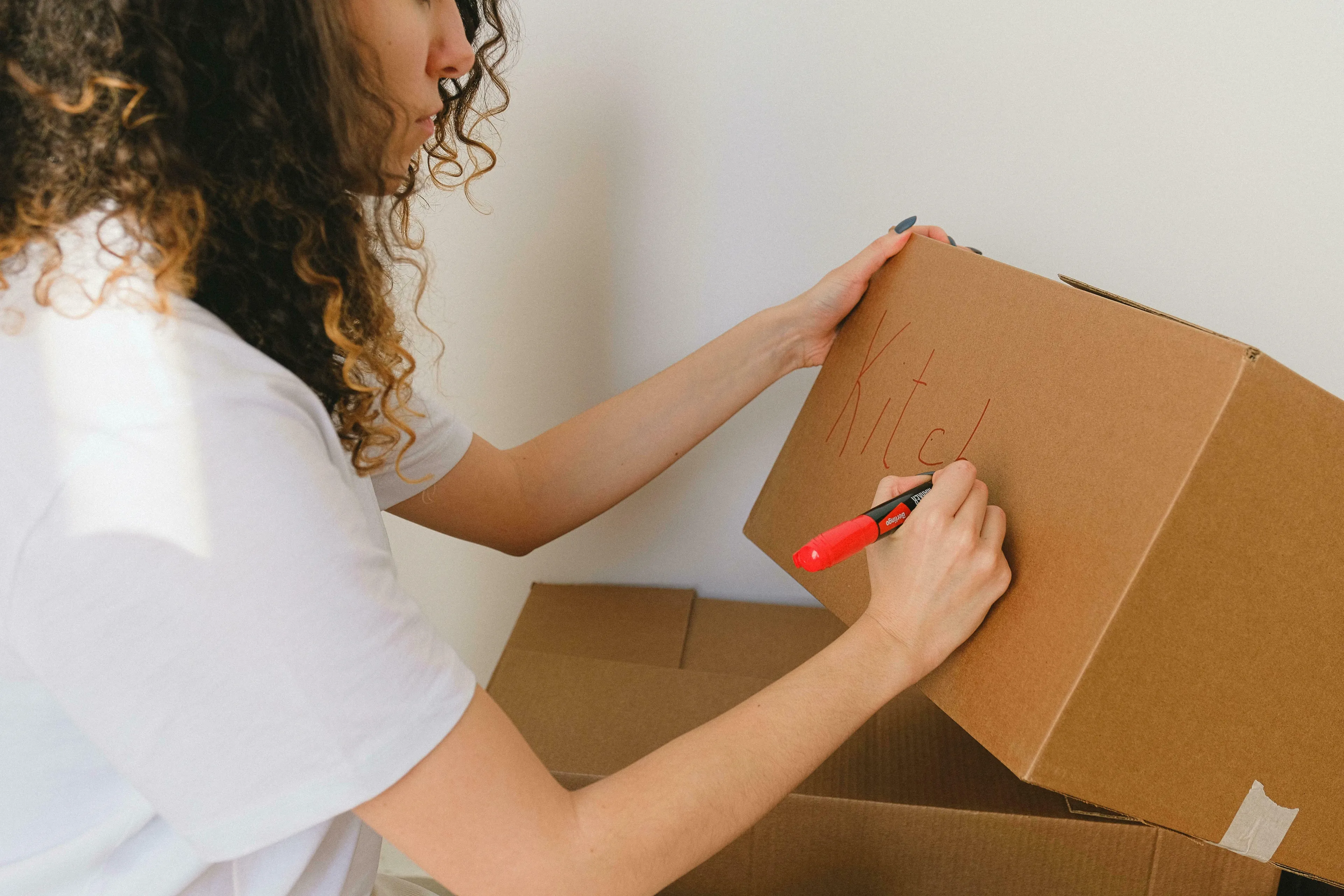 A woman with curly hair labels a moving box "Kitchen" with a red marker, reacting to change with proactive packing.