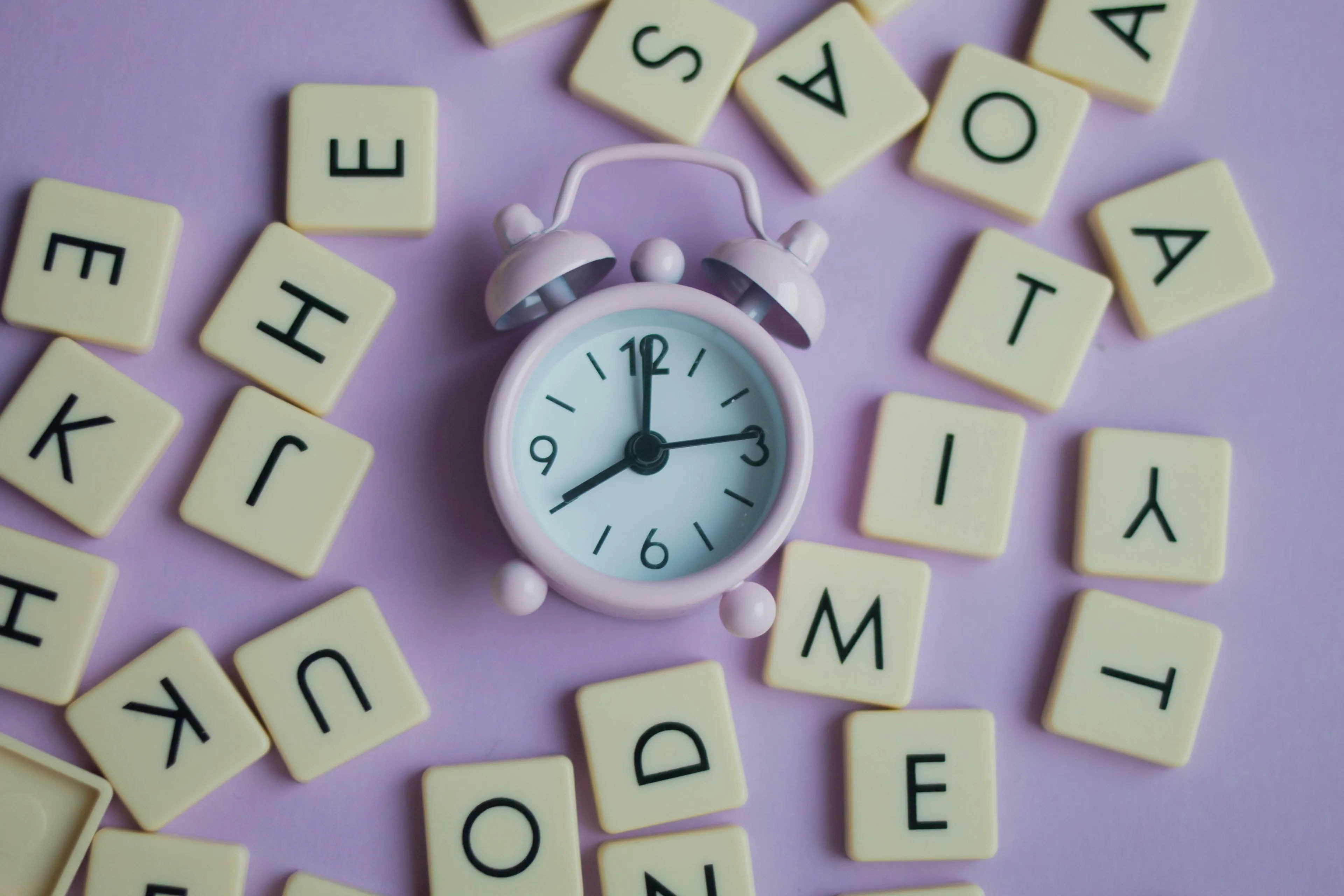 A pink alarm clock surrounded by letter tiles on a purple background, symbolizing time and routine. How do you react to change?