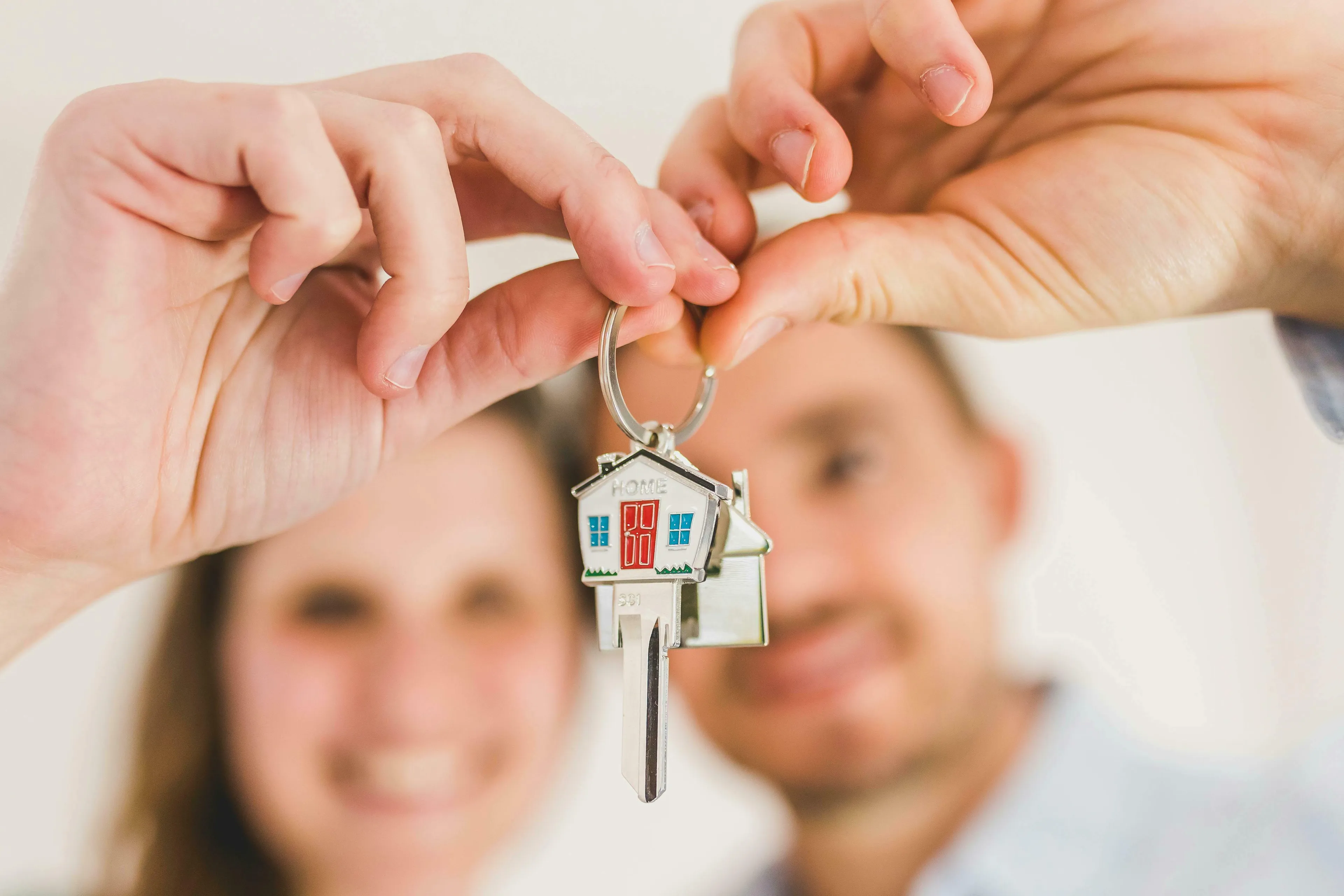 Hands holding new house keys, with a smiling couple blurred in the background. React to change.