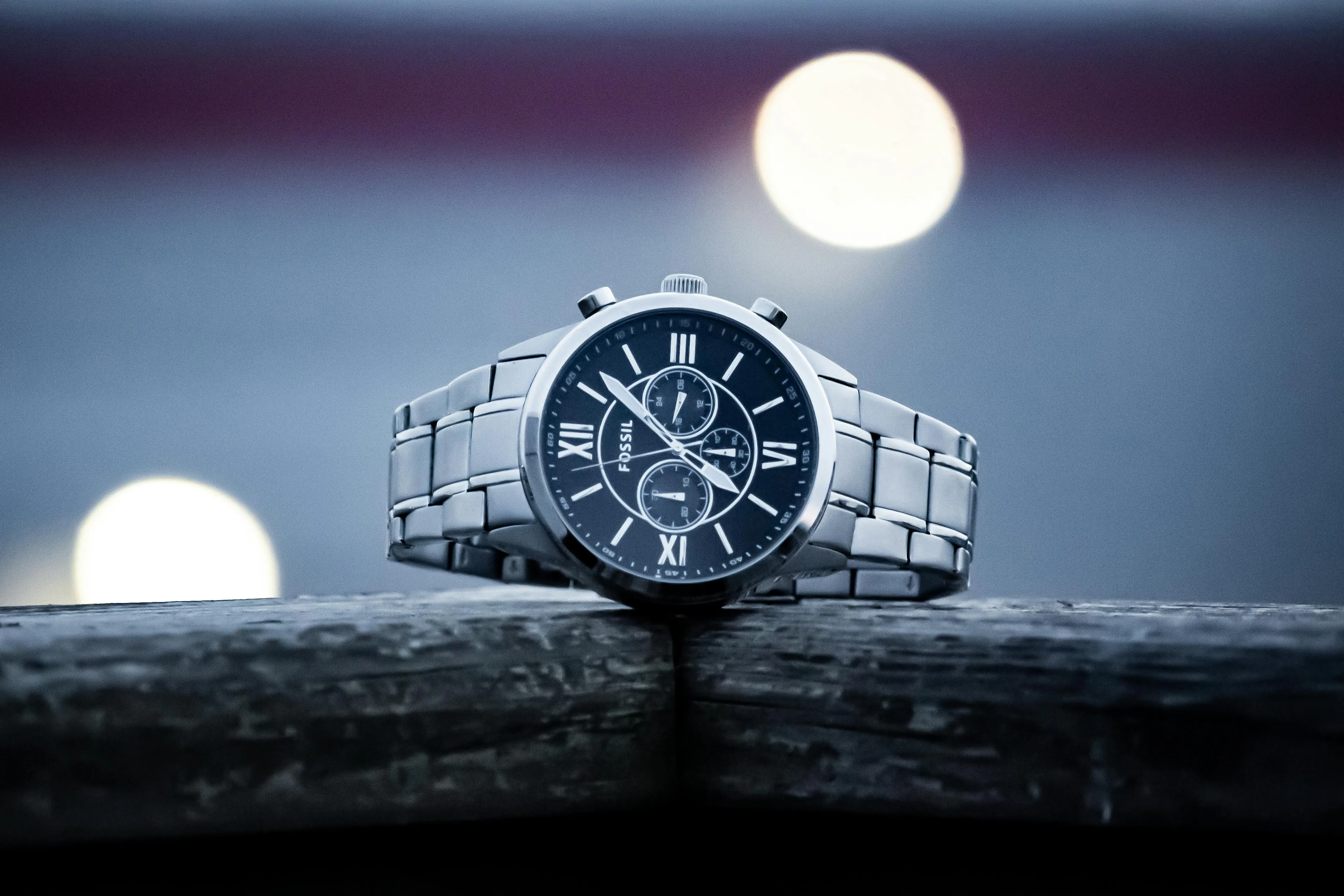 A silver Fossil watch with a dark face and Roman numerals, resting on a wooden surface with bokeh lights in the background.