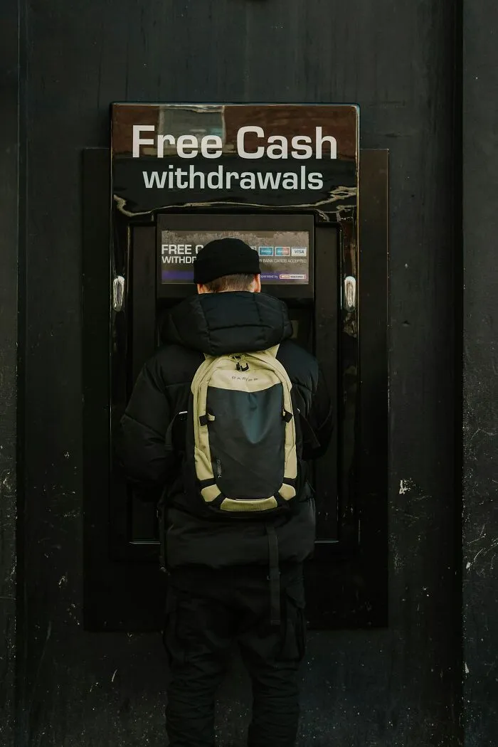 A person in a black jacket and backpack uses an ATM displaying Free Cash withdrawals. Visiting the US moments.