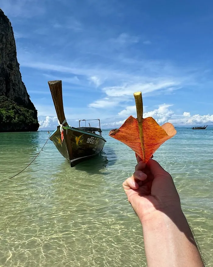 A hand holds a dry leaf, creatively manipulated to resemble a boat against a clear blue sky, a boat in the distance. Photo manipulations make simple objects parts of cities.