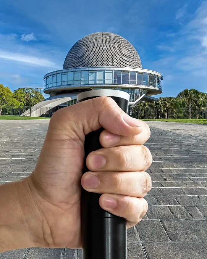 A hand manipulating a simple object, a microphone, to look like a part of a city with a planetarium in the background.