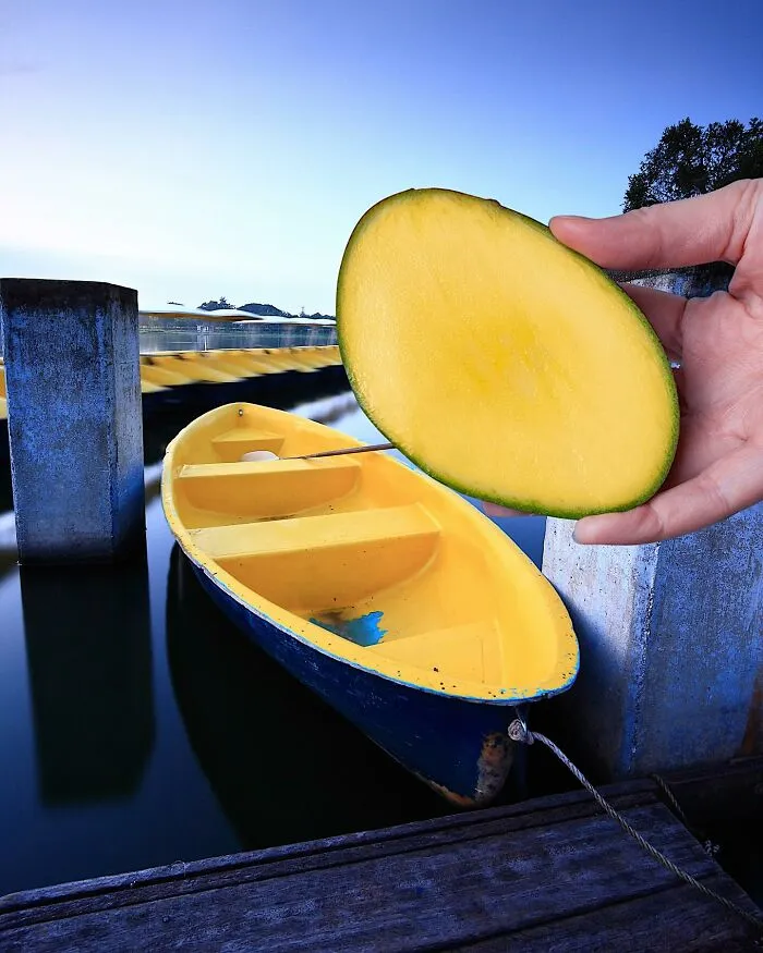 Clever photo manipulations: a hand holding a mango slice as if it's a boat in a small city harbor.